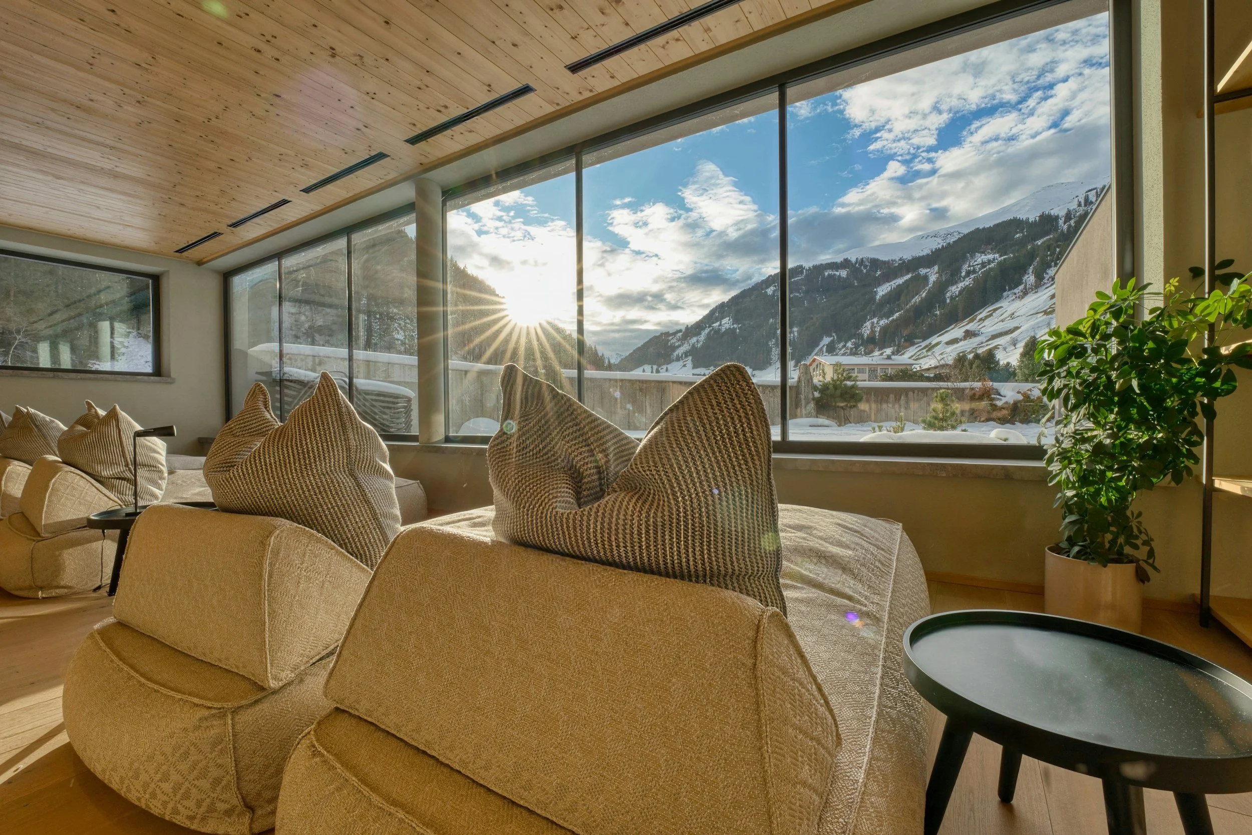 Living room with large glass windows showing snowy mountains and blue sky, beige sofa with pillows, black side table, and a green potted plant.