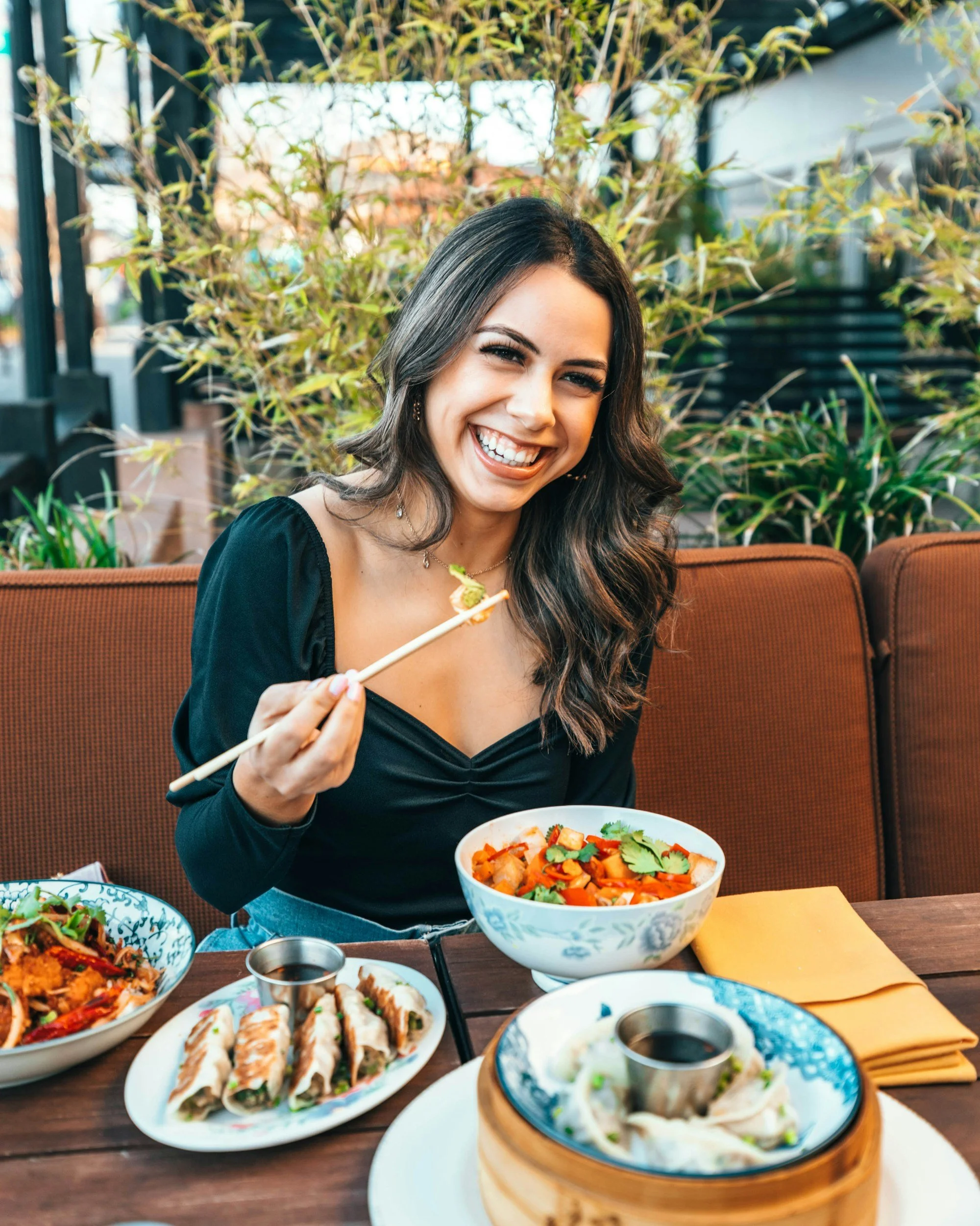 A woman enjoying an Asian meal outdoors, holding chopsticks, with various dishes including sushi, dumplings, and noodles on the table in front of her, colorful plants in the background.