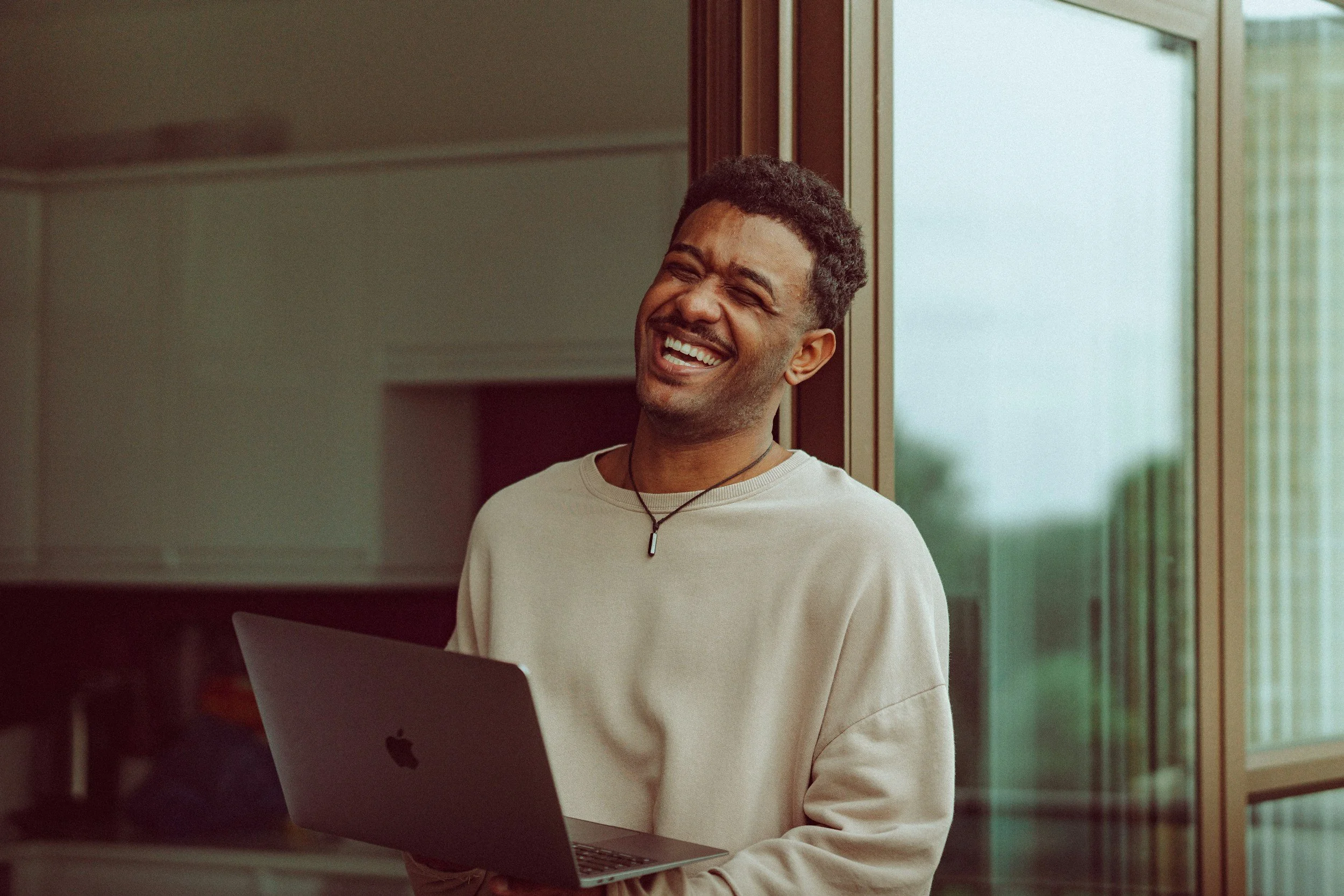 A young man with curly hair, wearing a white shirt and a necklace, smiling while sitting next to a window with a MacBook laptop in front of him.