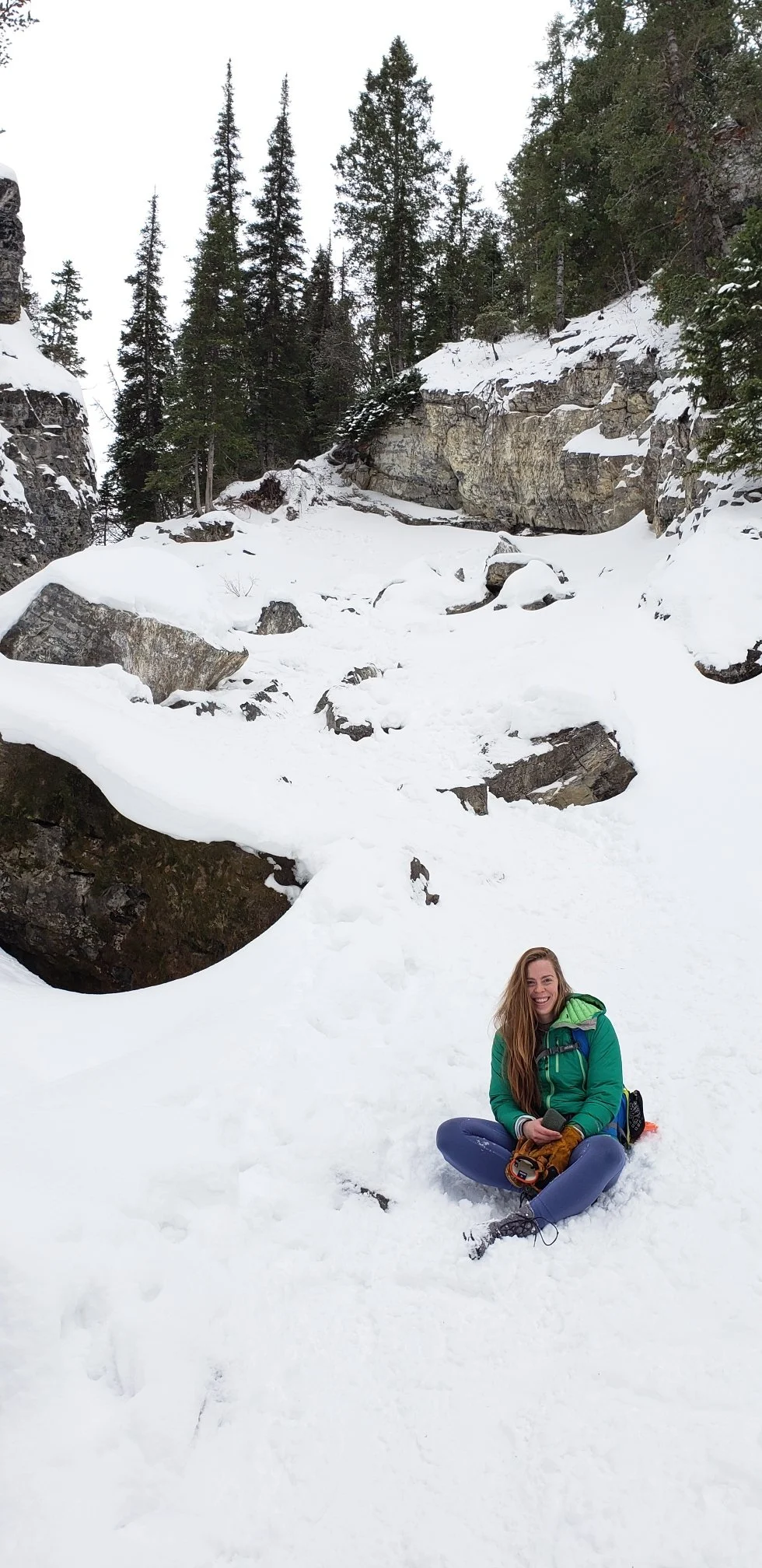 A smiling woman sitting on snow in a mountain valley with snow-covered rocks and evergreen trees.