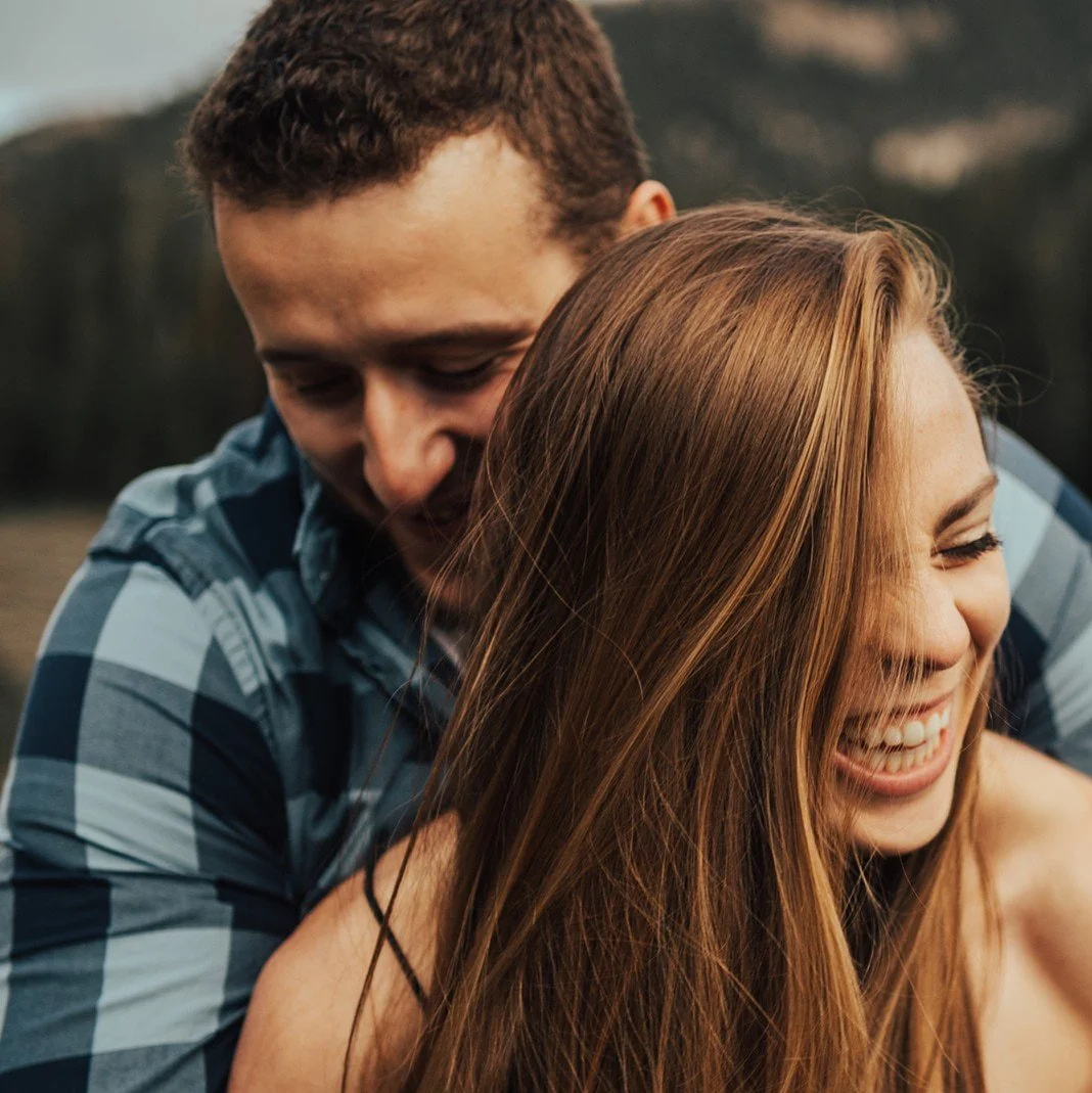 A couple sharing a joyful moment outdoors, with the woman smiling broadly and the man hugging her from behind.