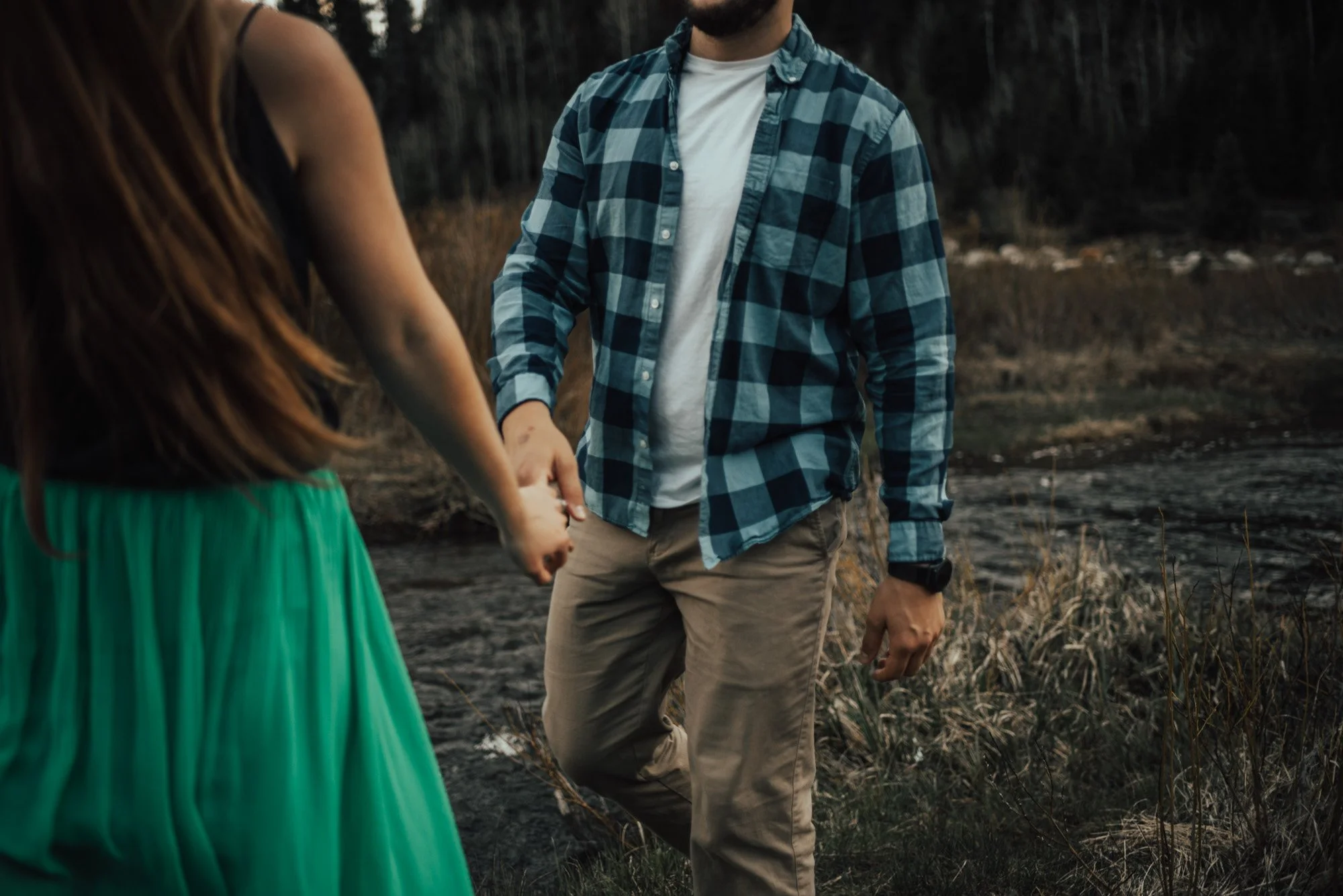 A man and a woman holding hands outdoors near a body of water with trees in the background. The woman is wearing a black top and a green skirt, and the man is wearing a blue plaid shirt over a white T-shirt and beige pants.