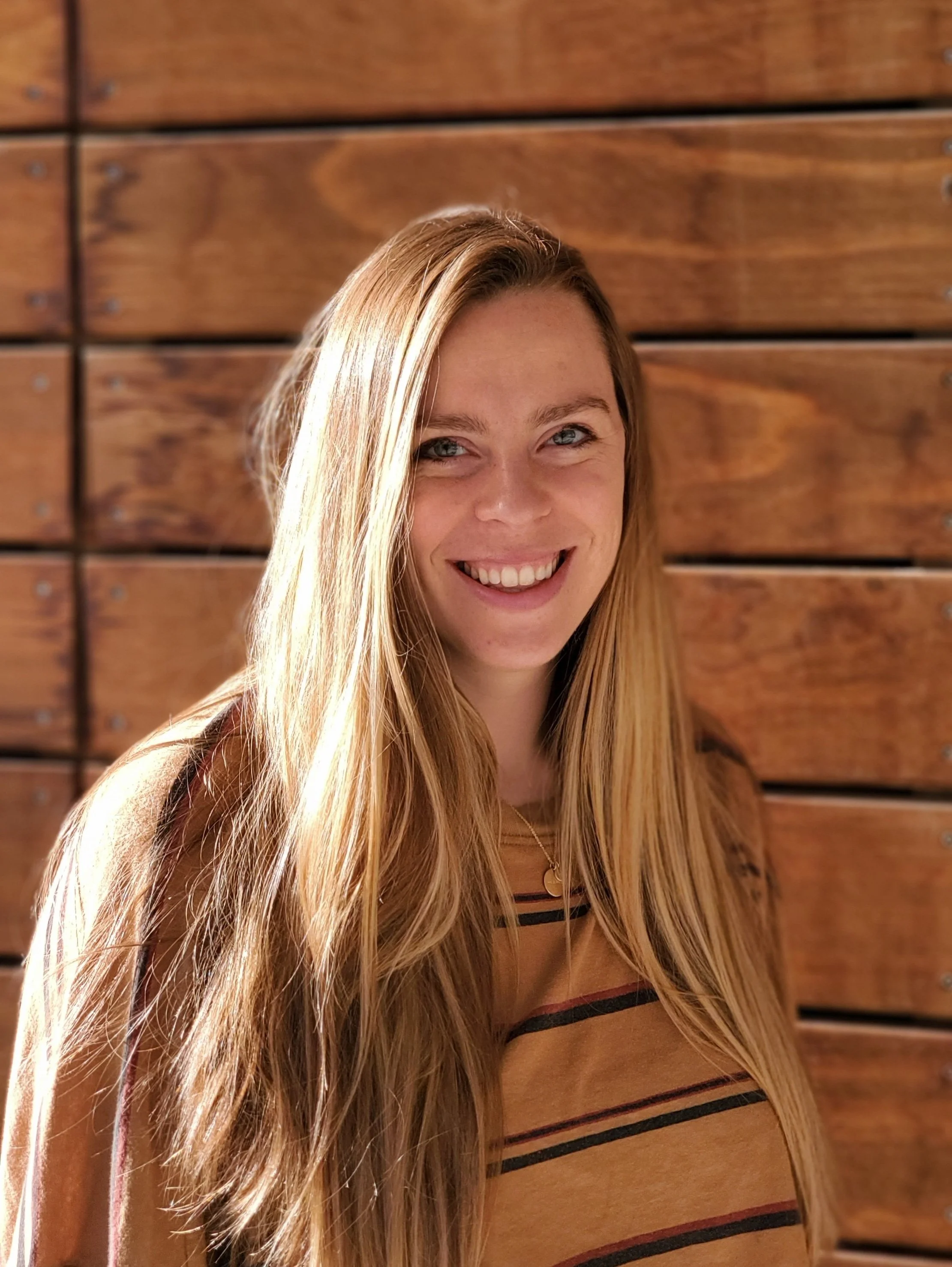 Alli smiling, standing in front of a wooden wall at the University of Utah where she completed her masters.