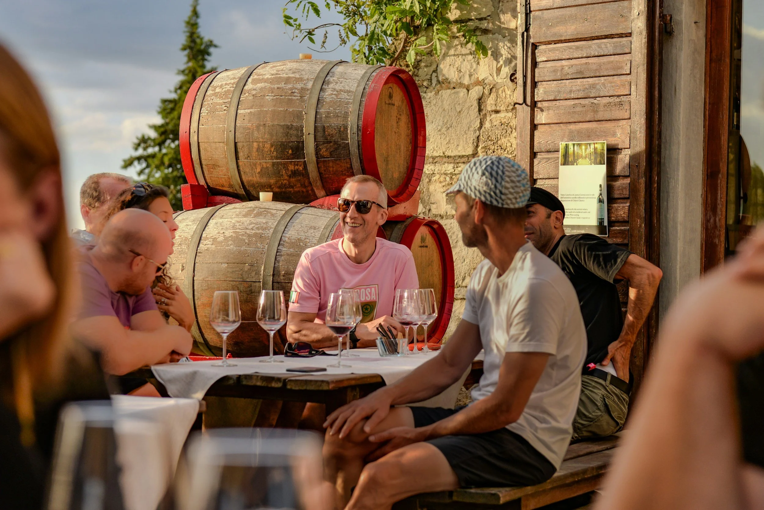 Man in pink shirt with sunglasses on in front of wine barrels smiling table with 7 almost empty glasses of red wine and a man in a white tshirt next to him
