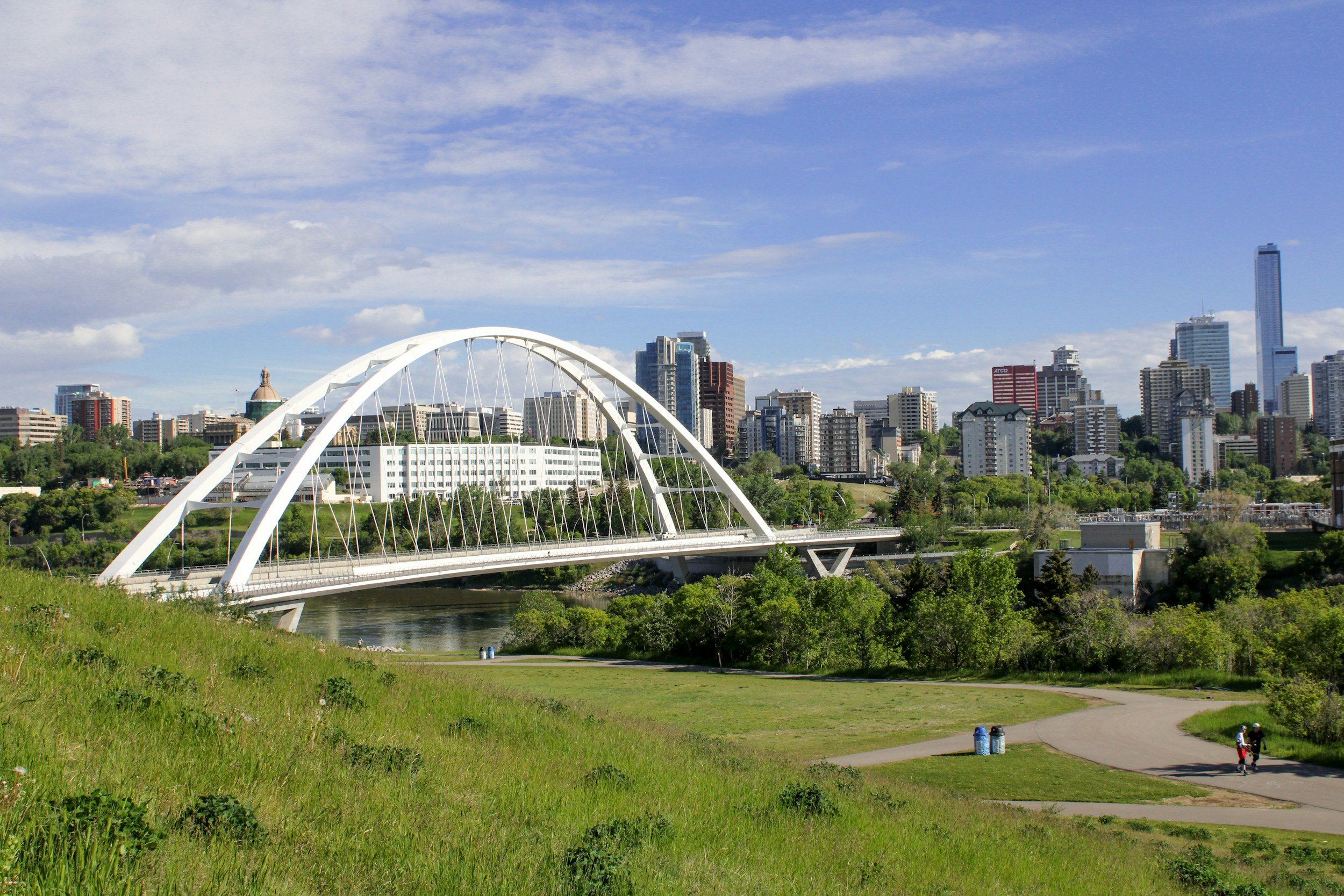 A city skyline with tall buildings and skyscrapers viewed from a park with a bridge crossing a river, under a partly cloudy sky.