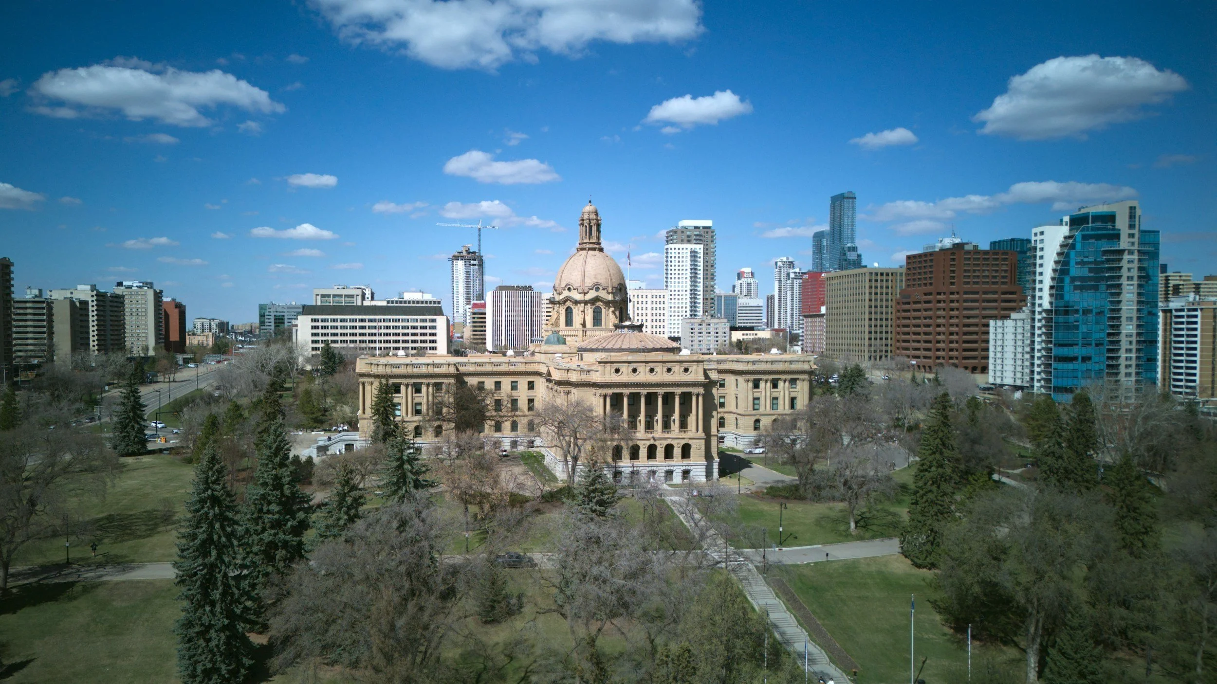A city skyline with a historic government building in the foreground, surrounded by trees and green spaces, and modern skyscrapers in the background under a blue sky with scattered clouds.
