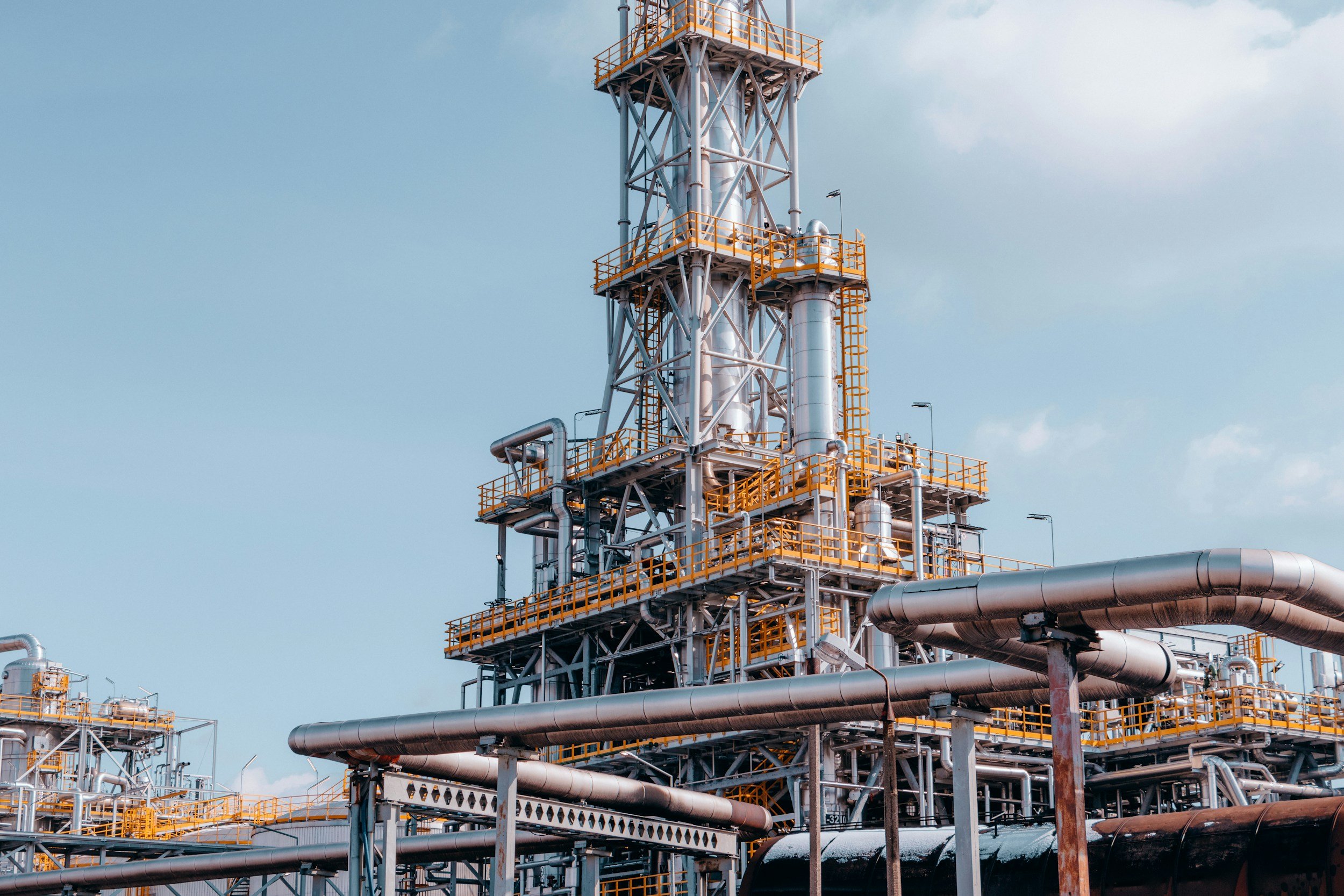 Industrial refinery with metal pipes, orange safety railings, and a tall metal structure under a partly cloudy sky.