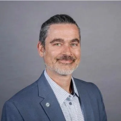 A man with gray hair and beard wearing a blue blazer and white patterned shirt, smiling at the camera against a gray background.