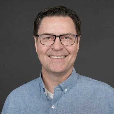 A man with short dark hair, glasses, and a light blue button-up shirt smiling at the camera.