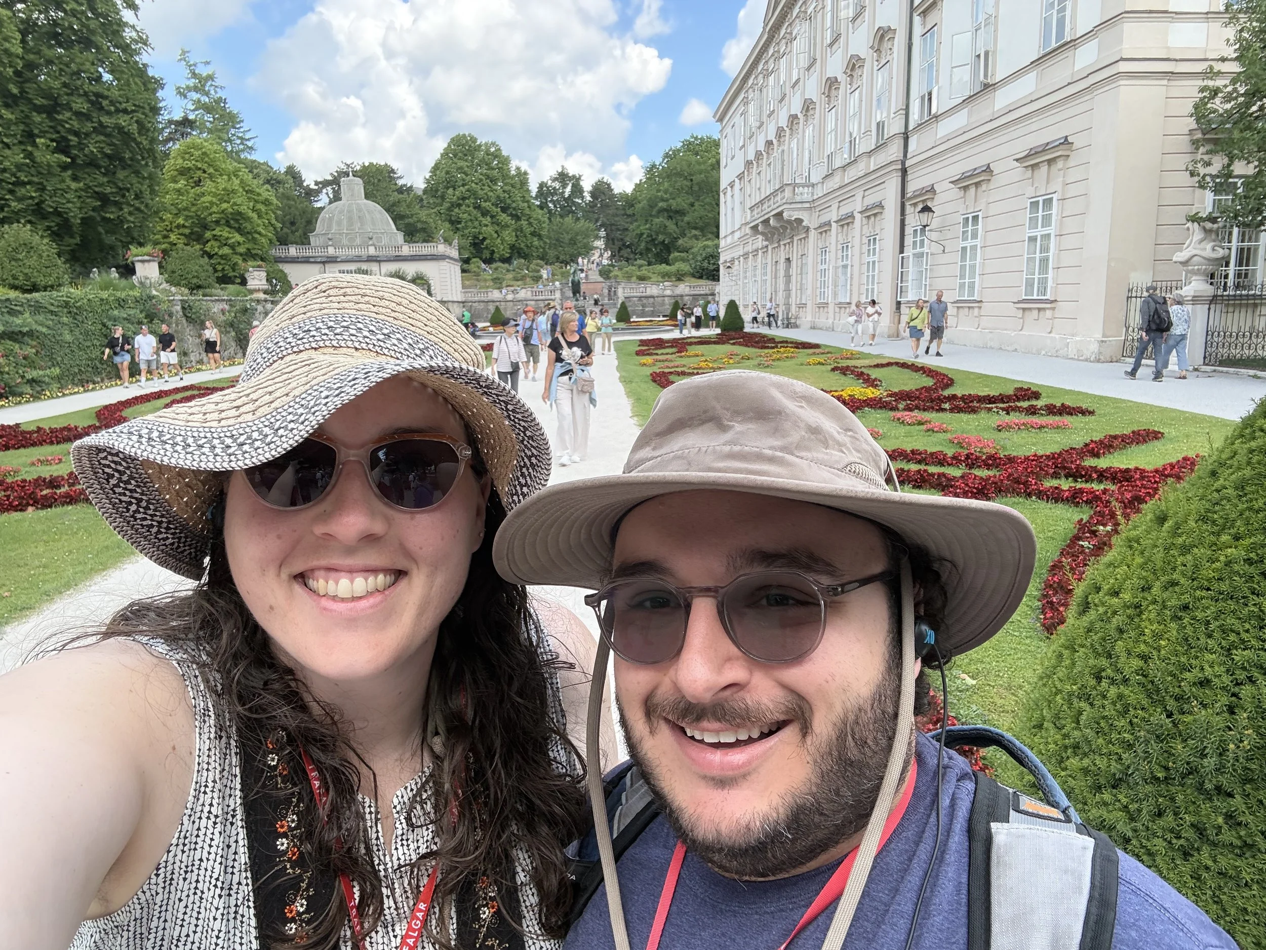 Two smiling tourists taking a selfie in front of a historic building with a well-maintained garden, many other visitors walking around on a bright, partly cloudy day.