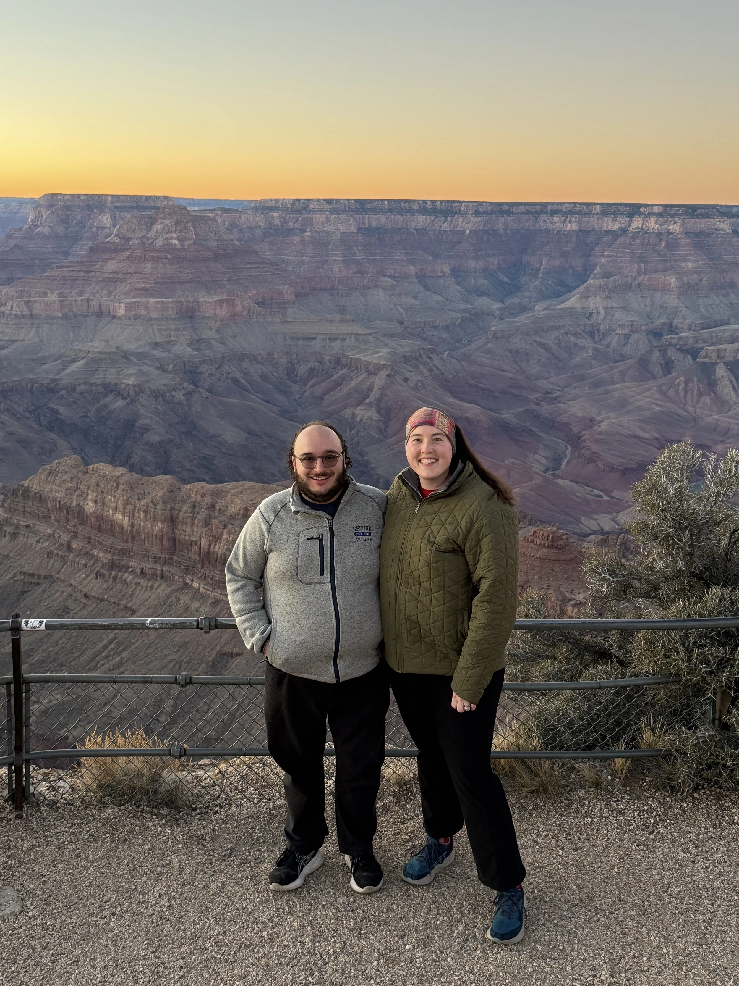 Two smiling people standing in front of the Grand Canyon during sunset, with a metal fence and desert shrubs in the foreground.