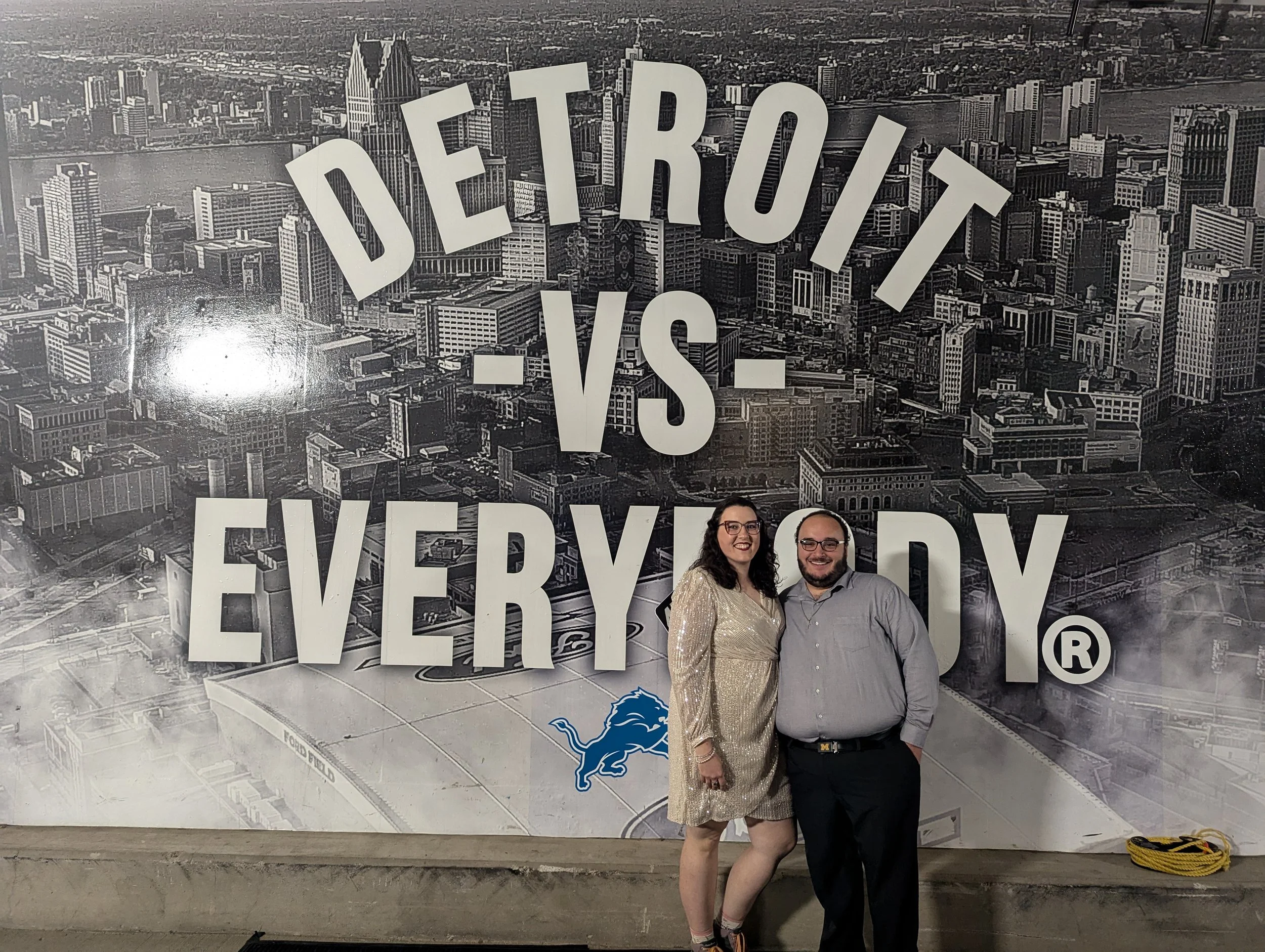 Two people standing in front of a black and white cityscape background with large white text that reads "DETROIT VS. EVERBODY". The woman is wearing a sparkly gold dress and glasses, while the man is wearing glasses and a gray shirt. They are smiling at the camera.