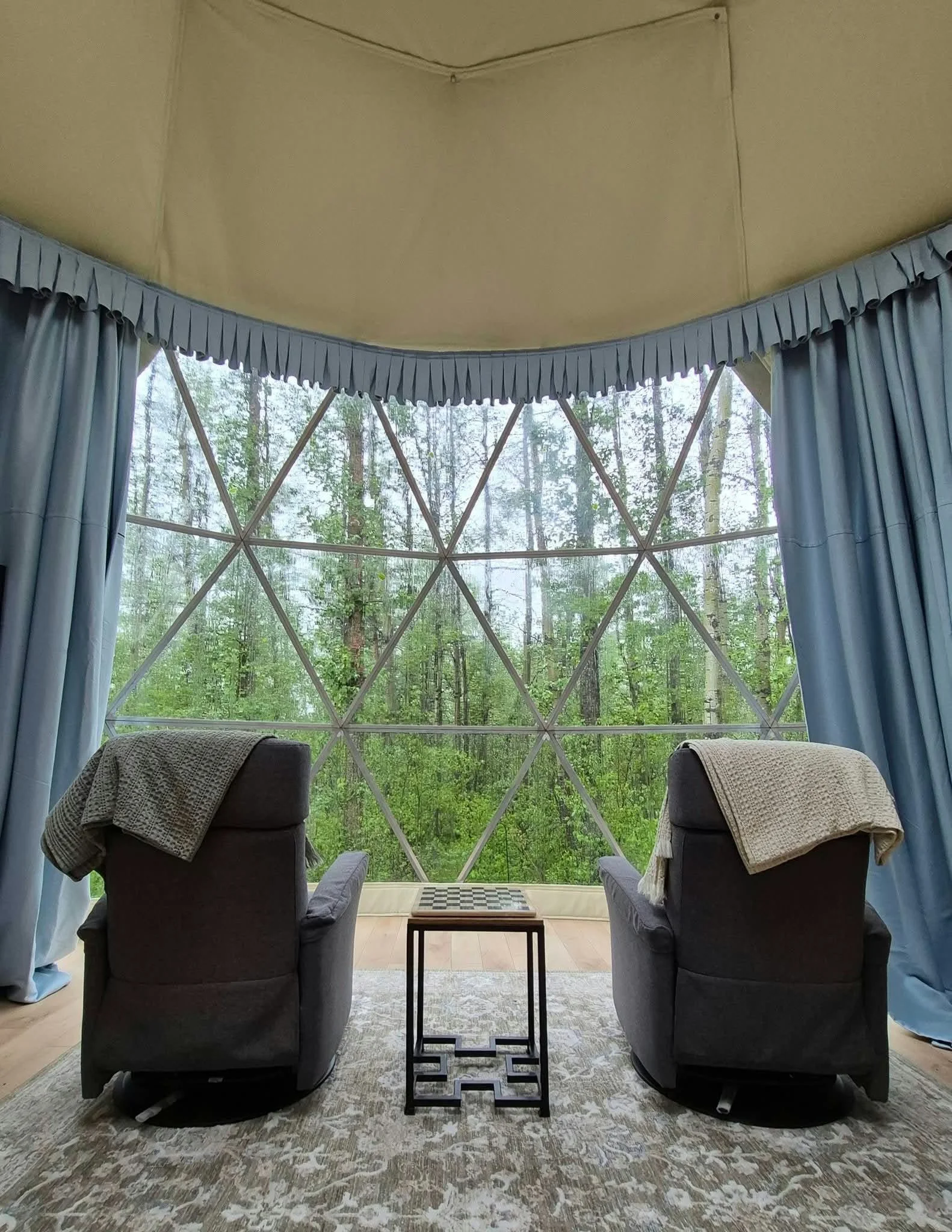 Inside view of dome with two armchair chairs, a coffee table, and a large window with a view of a green forest. Curtains frame the window, and the ceiling is beige with a corner cord that might control window blinds.