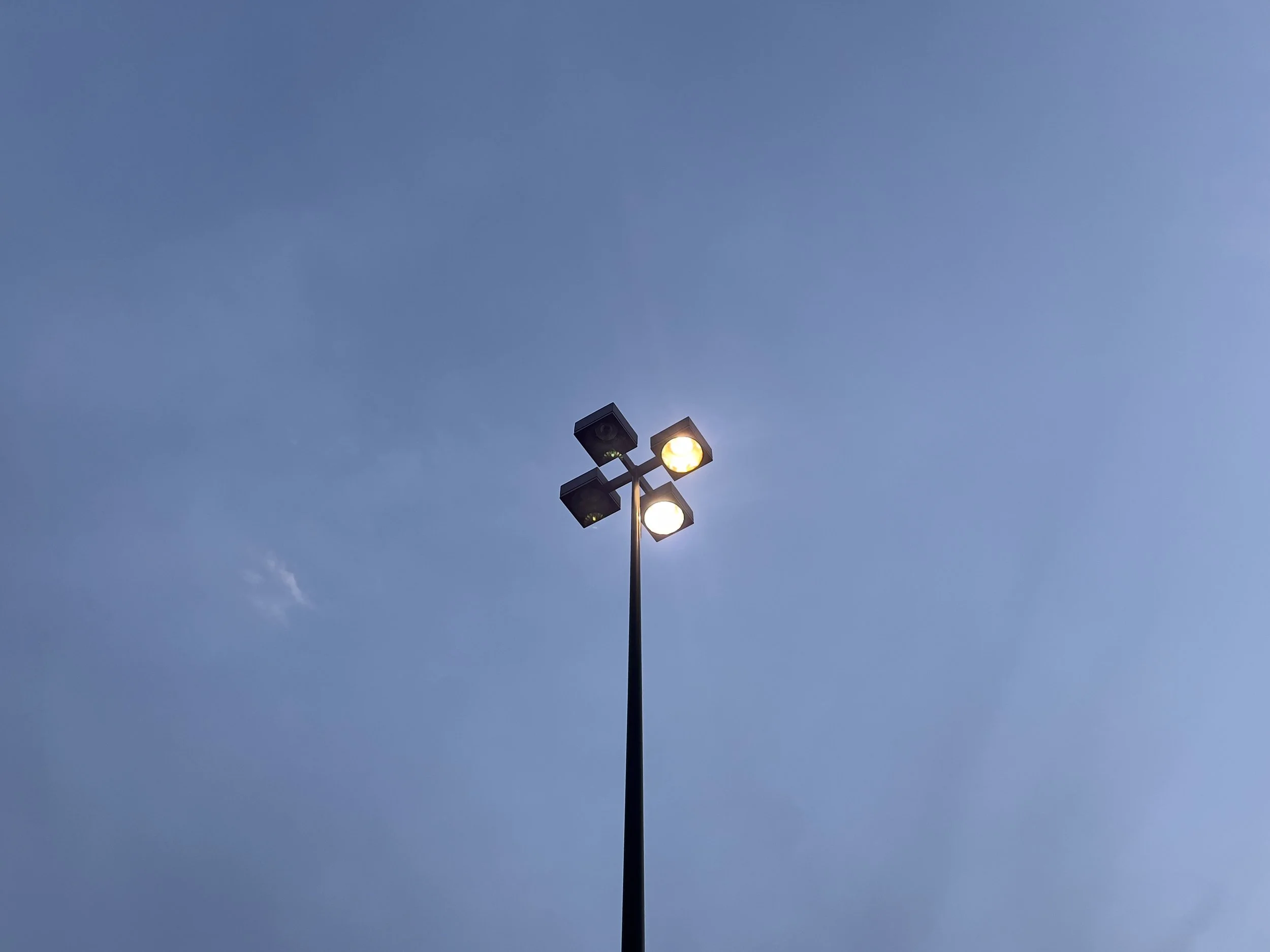 A tall streetlight with four lights against a dark blue sky near dusk, with only two of the lights working