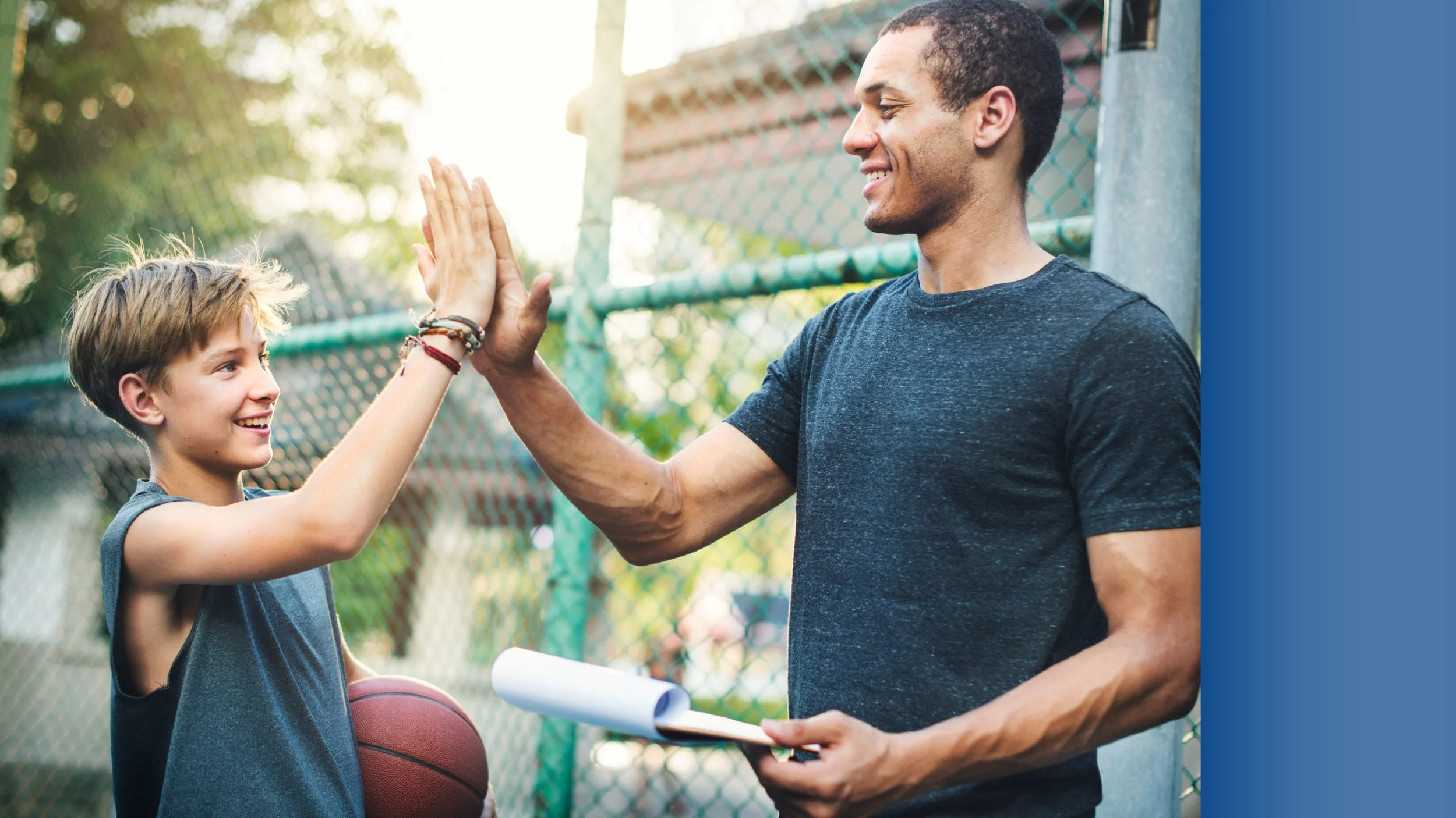A young boy in a sleeveless shirt high-fives an adult man at a basketball court. The boy is holding a basketball and smiling, while the man is smiling and holding a rolled-up paper and a clipboard. They are standing next to a chain-link fence with trees in the background on a sunny day.