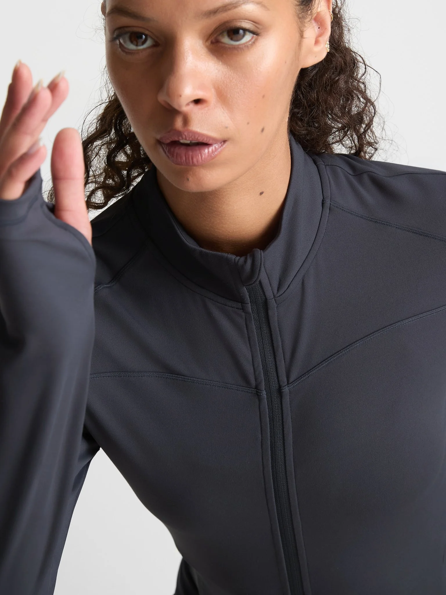 A woman with curly hair wearing a black athletic jacket, holding her left hand up near her face, with a neutral expression, against a plain light background.