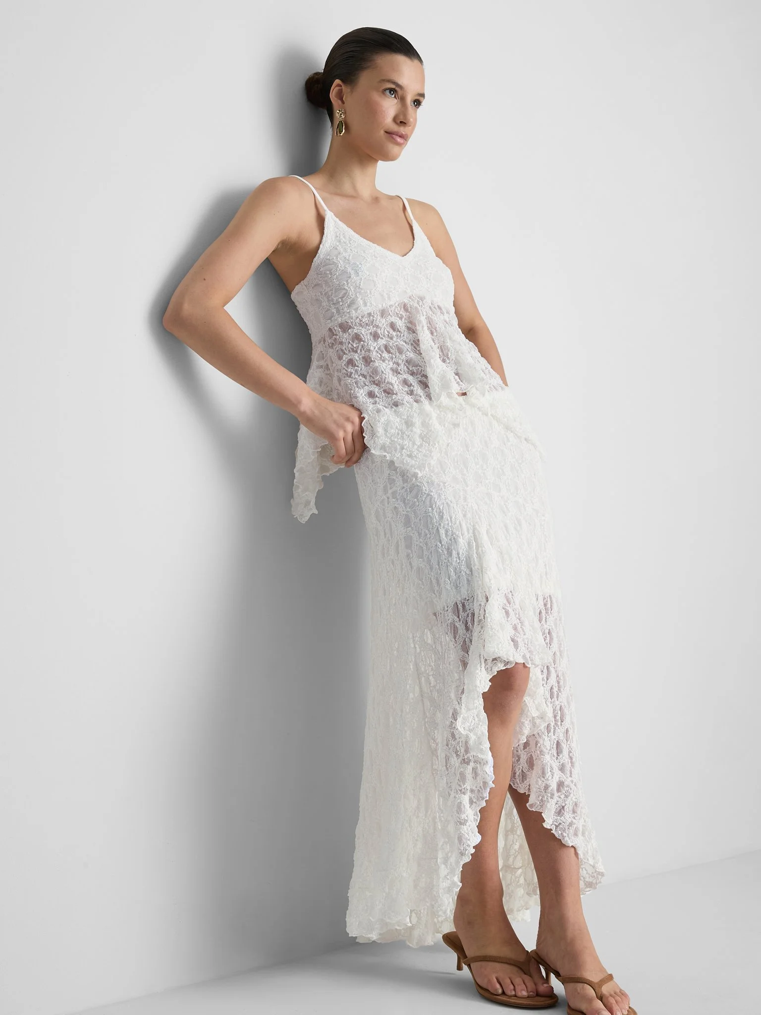 Woman in a white lace sleeveless dress, standing against a plain white wall, with her hands in her pockets, wearing earrings and brown sandals.