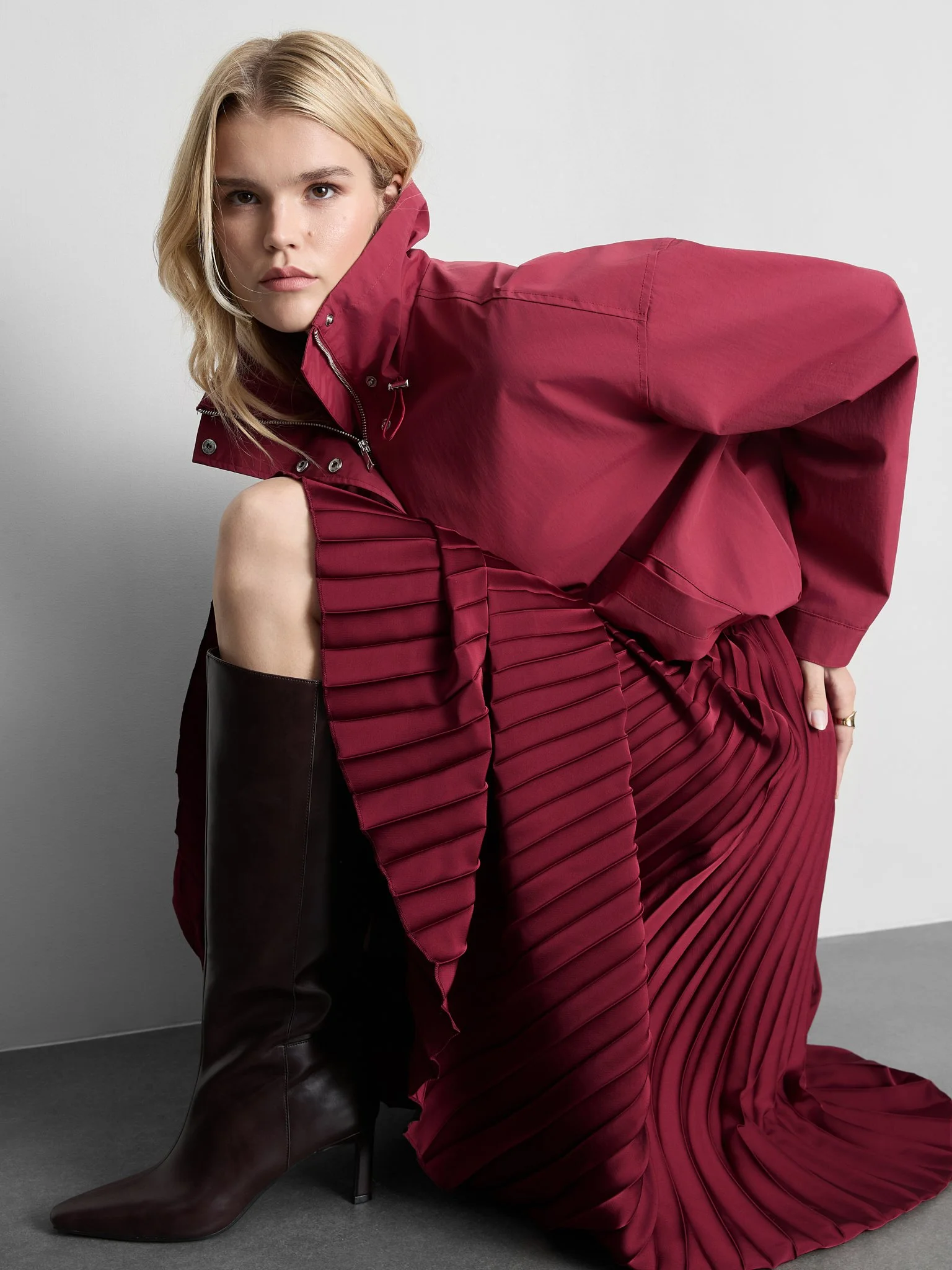 A woman wearing a red jacket and a matching red dress with pleated details, sitting on the floor with one knee slightly raised and wearing a dark brown knee-high boot.