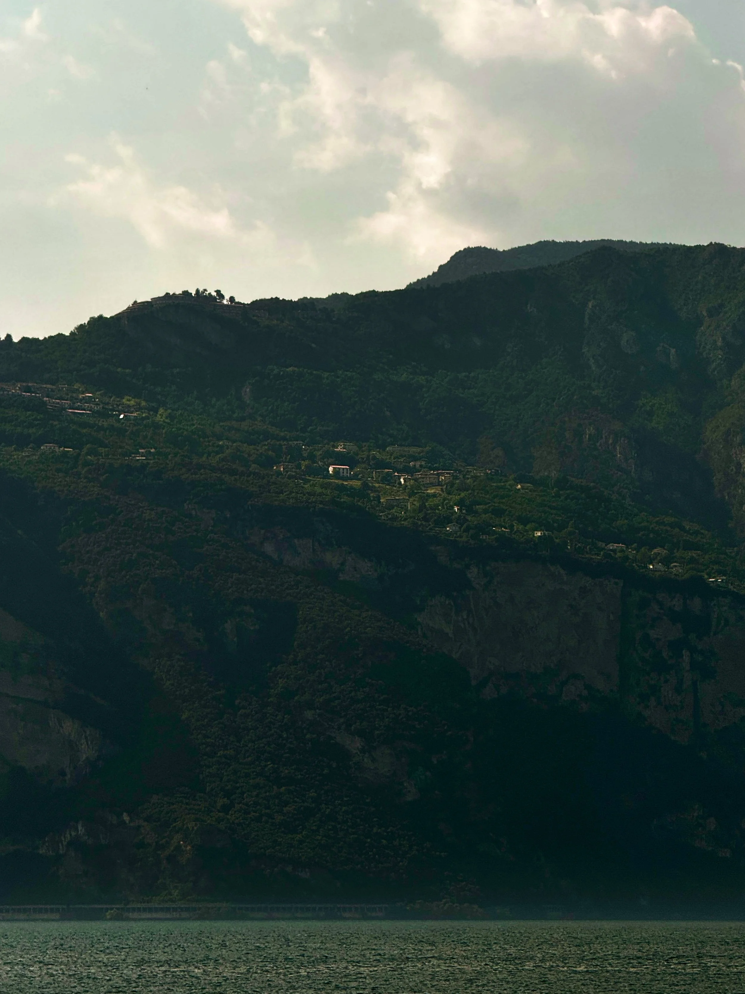 A scenic view of a mountain range with green hills and scattered trees, under a partly cloudy sky, overlooking fields in the foreground.