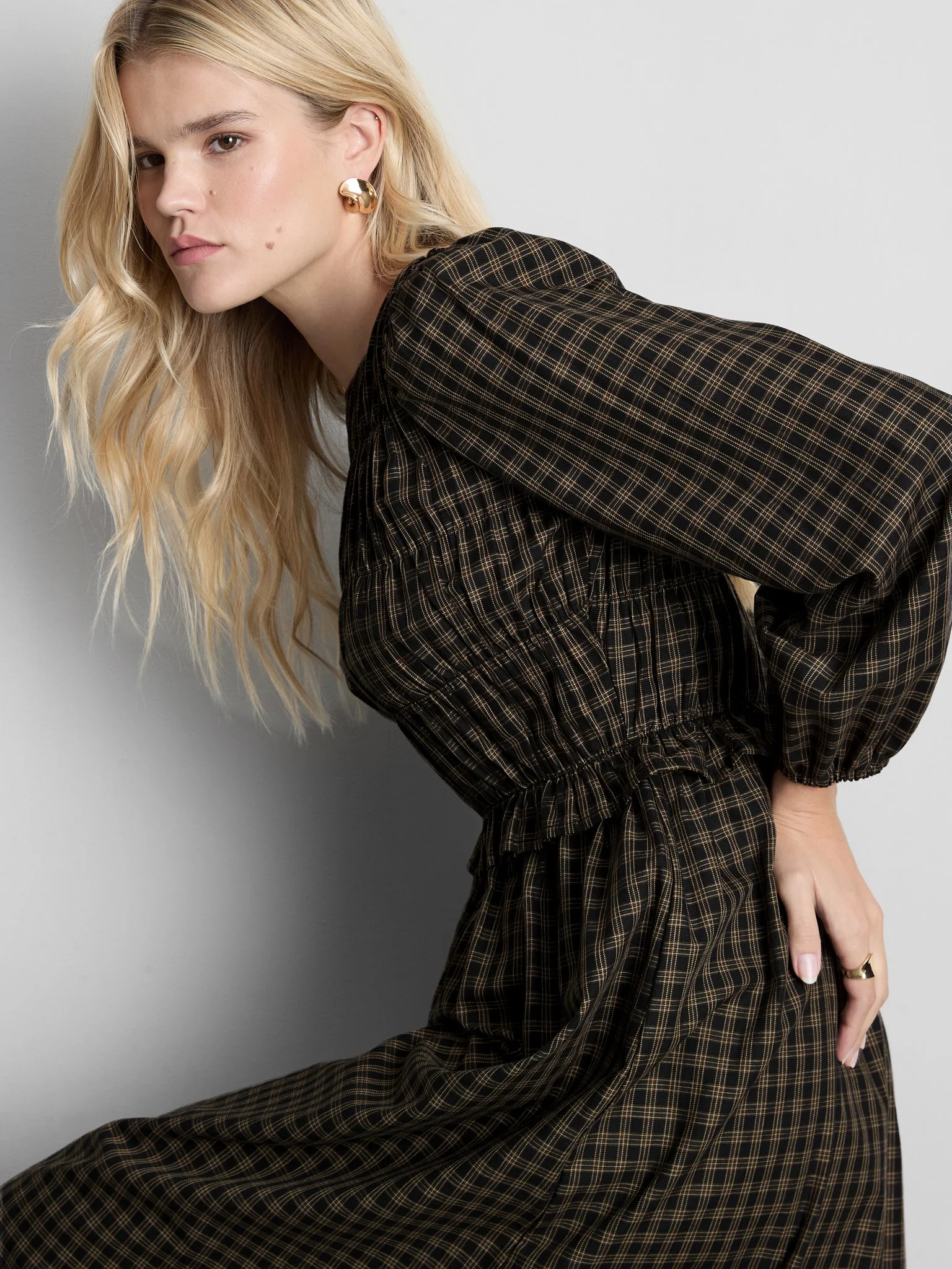 A woman with long blonde hair and fair skin, wearing a brown checked outfit, is sitting against a gray wall, looking directly at the camera with a serious expression.