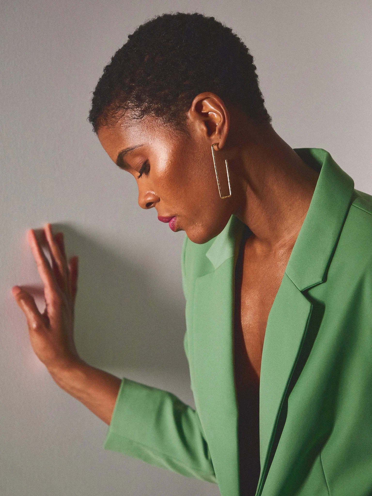 A woman with short curly hair and dark skin wearing a bright green blazer and rectangle-shaped gold earrings, posing with her eyes closed and face turned downward against a plain gray background.