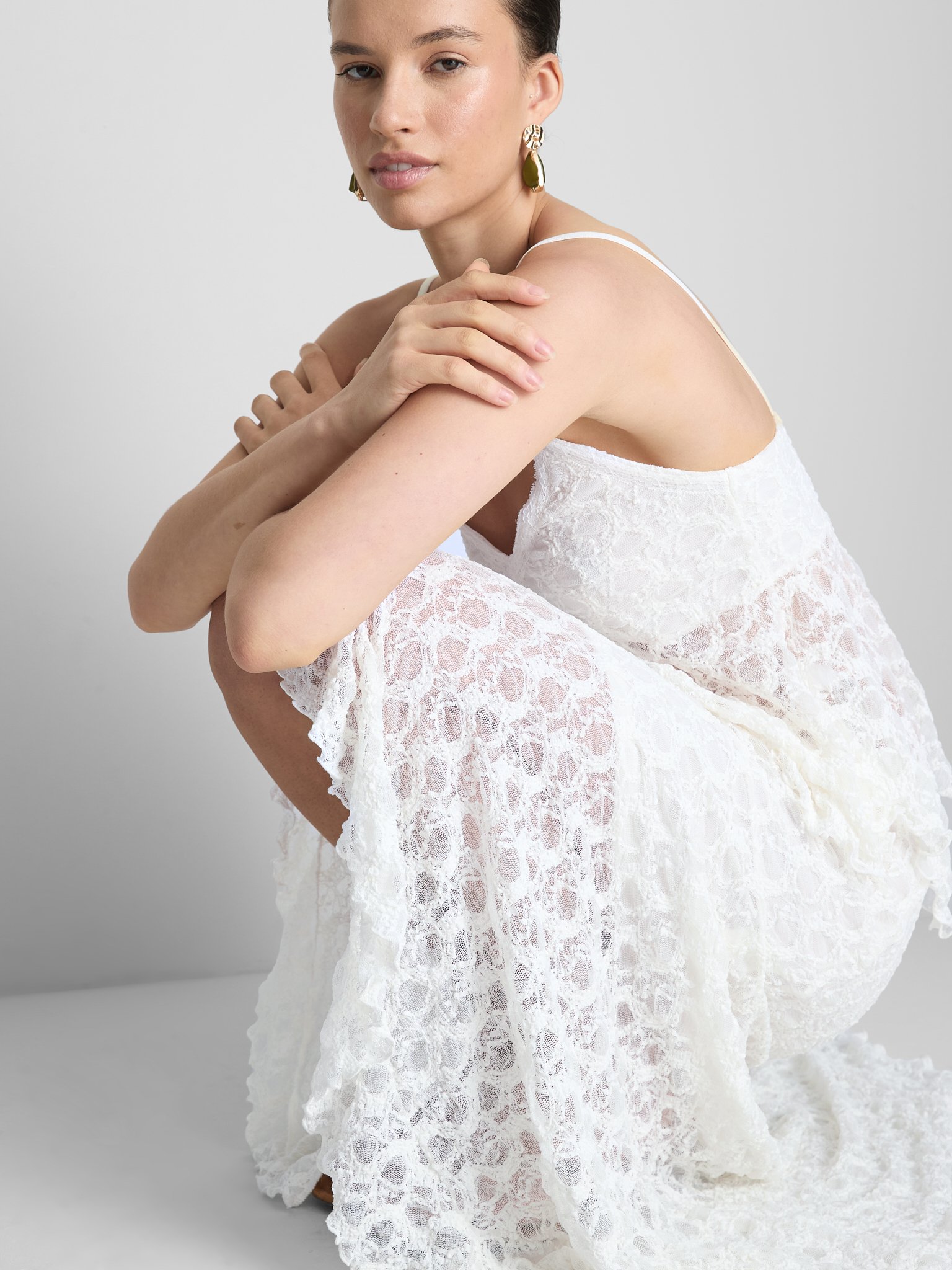 A woman in a white lace dress with spaghetti straps, sitting on the ground with her knees pulled close, resting her arms on her knees, against a plain gray background. She has short dark hair, wears large gold and dark green earrings, and is looking 