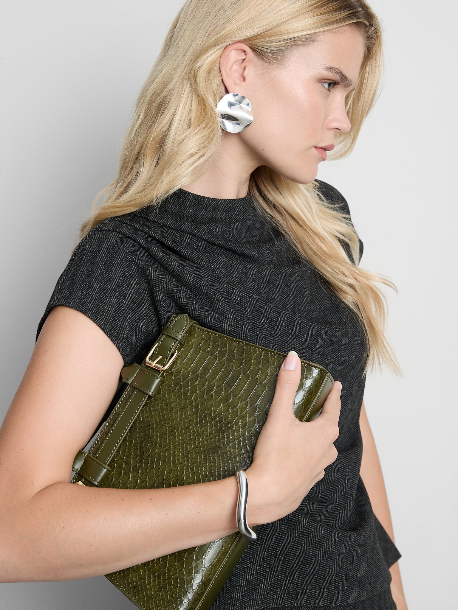 A woman with blonde hair wearing a black textured dress and large silver earrings, holding an olive green crocodile-patterned clutch purse against a light gray background.