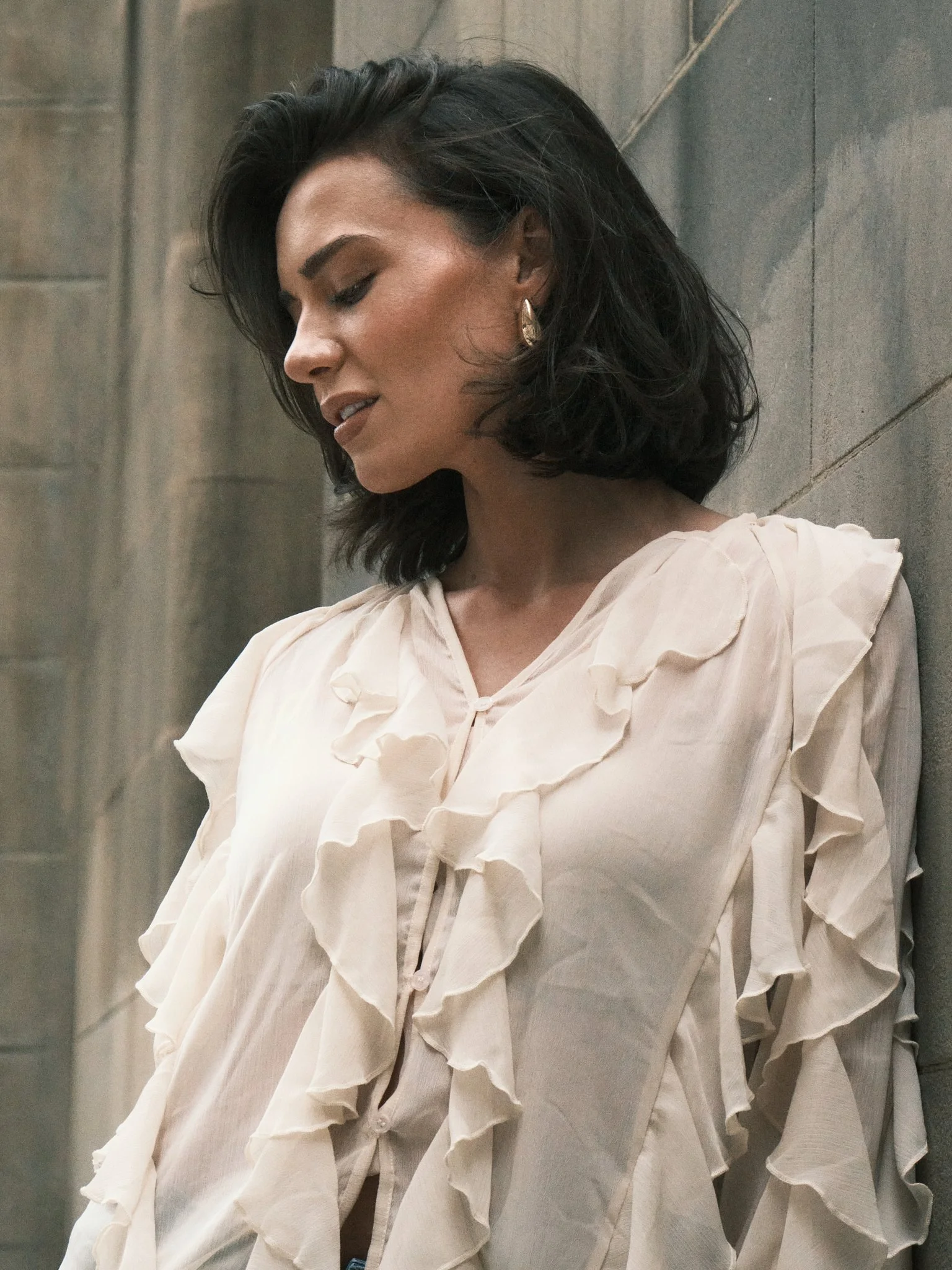 A woman with dark, wavy hair and a light cream-colored blouse with ruffled details standing against a gray stone wall, looking down.