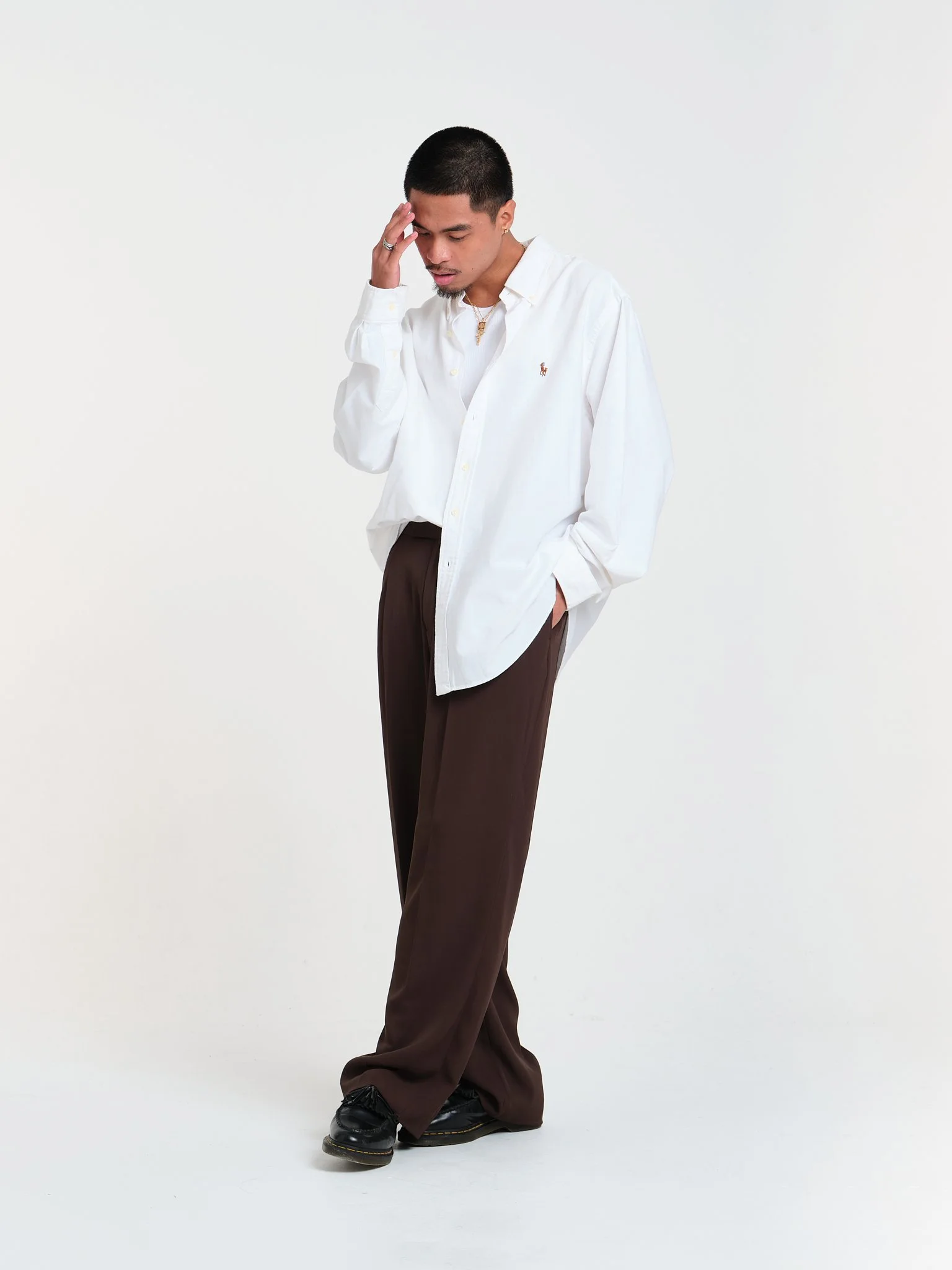 Young man wearing a white button-down shirt, brown pants, and black shoes, standing against a plain light background with one hand touching his forehead, looking down.