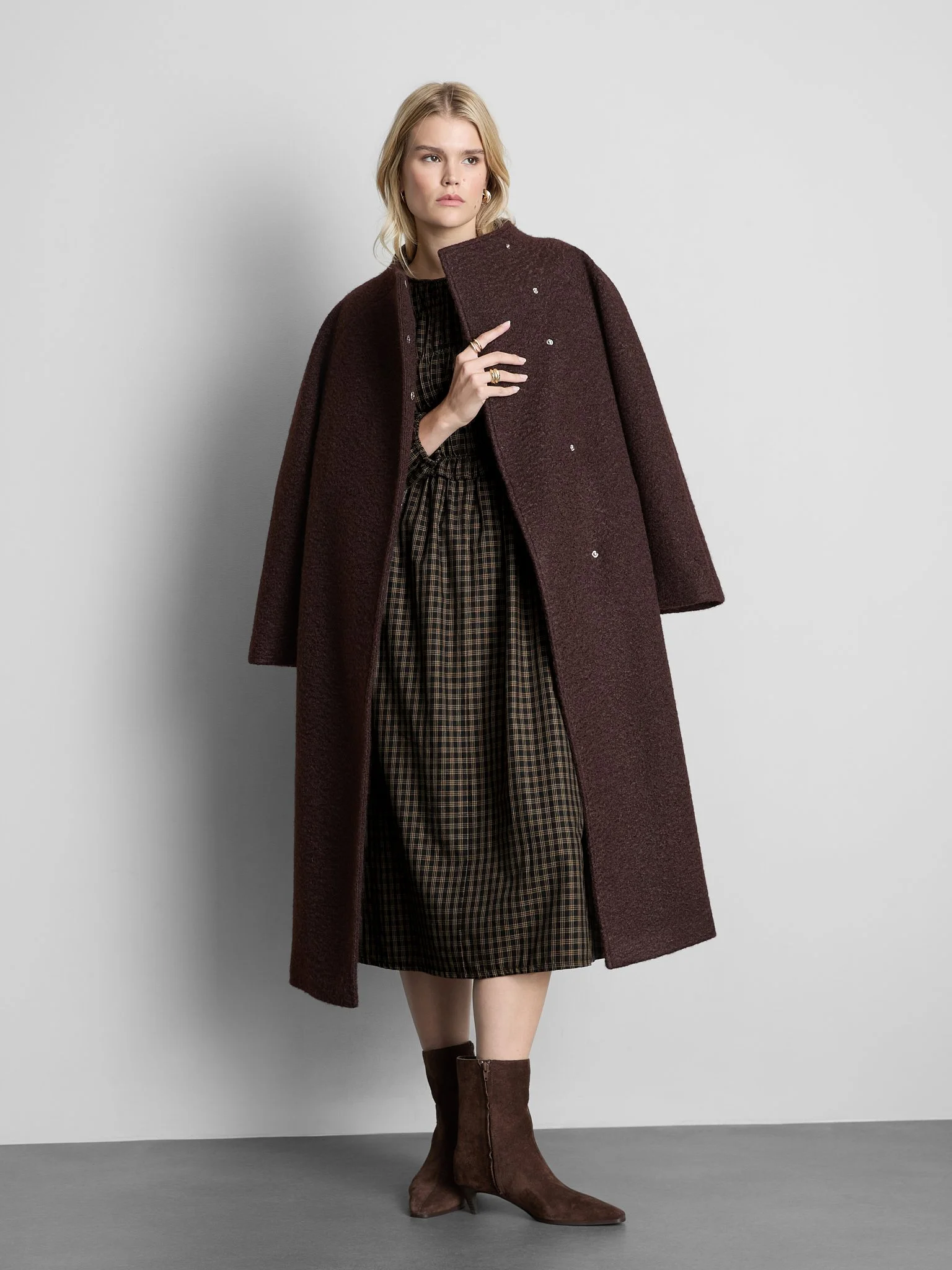 A young woman with blonde hair wearing a brown coat, plaid dress, and brown boots standing against a plain light gray background.