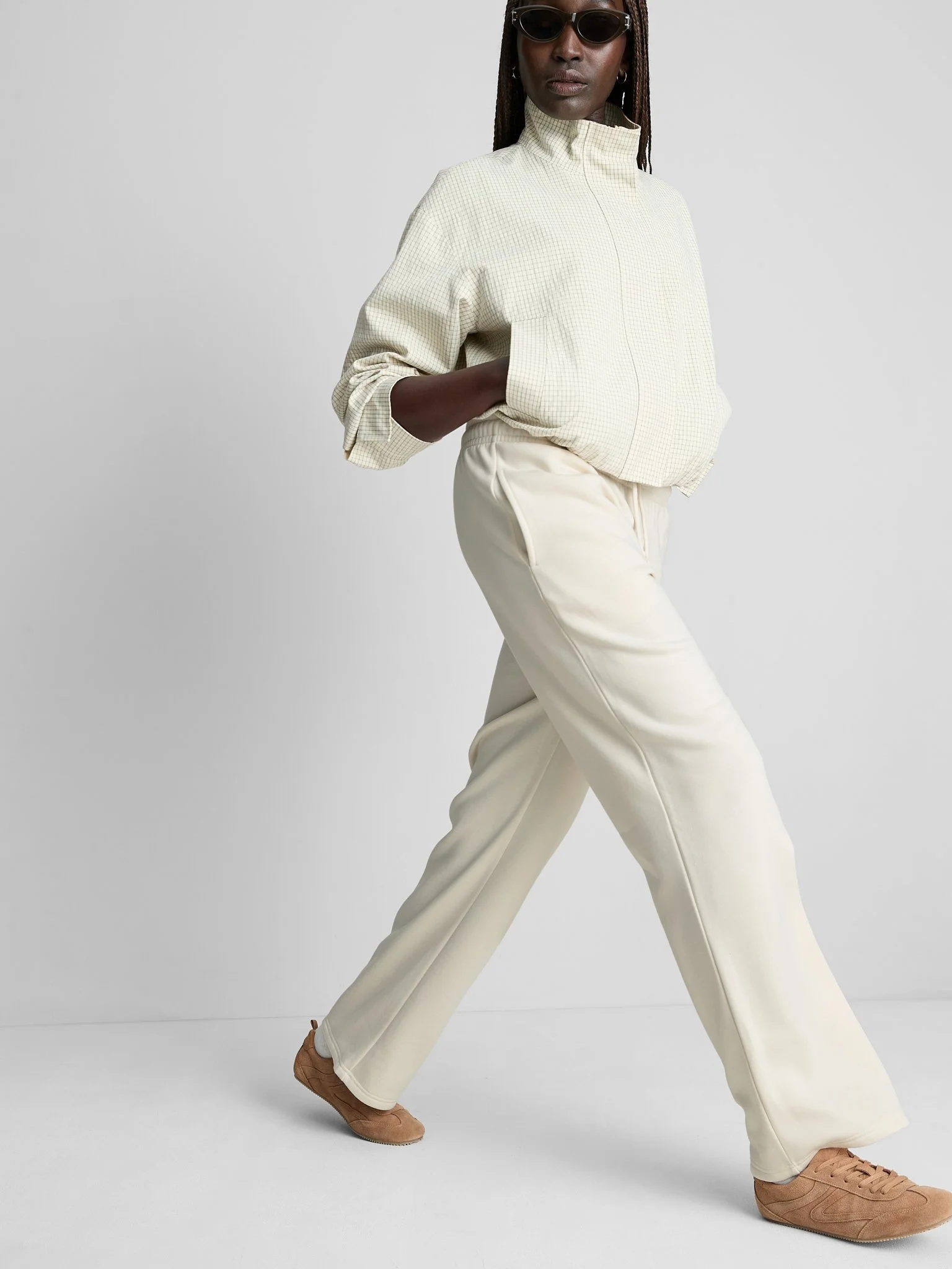 A woman in casual beige and cream-colored clothing, with sunglasses and brown sneakers, standing against a plain white background.