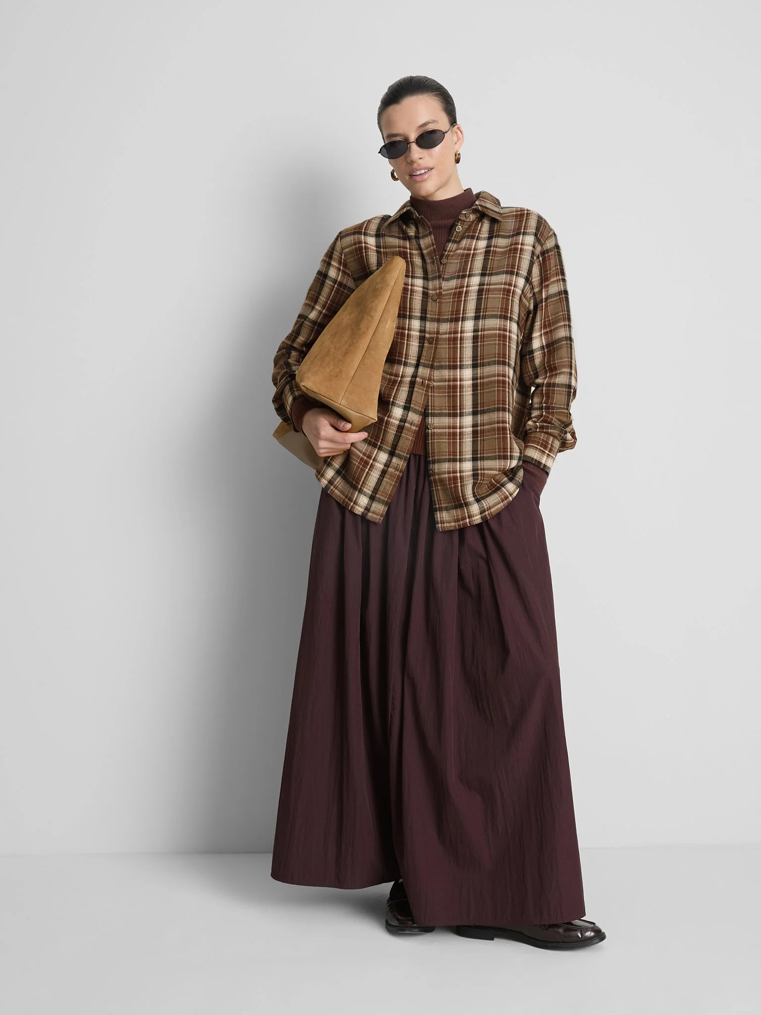 A woman wearing a plaid button-up shirt over a dark turtleneck, wide-legged maroon pants, and black shoes, holding a tan tote bag. She has dark hair slicked back and is wearing black oval sunglasses and earrings, standing against a plain gray backgro