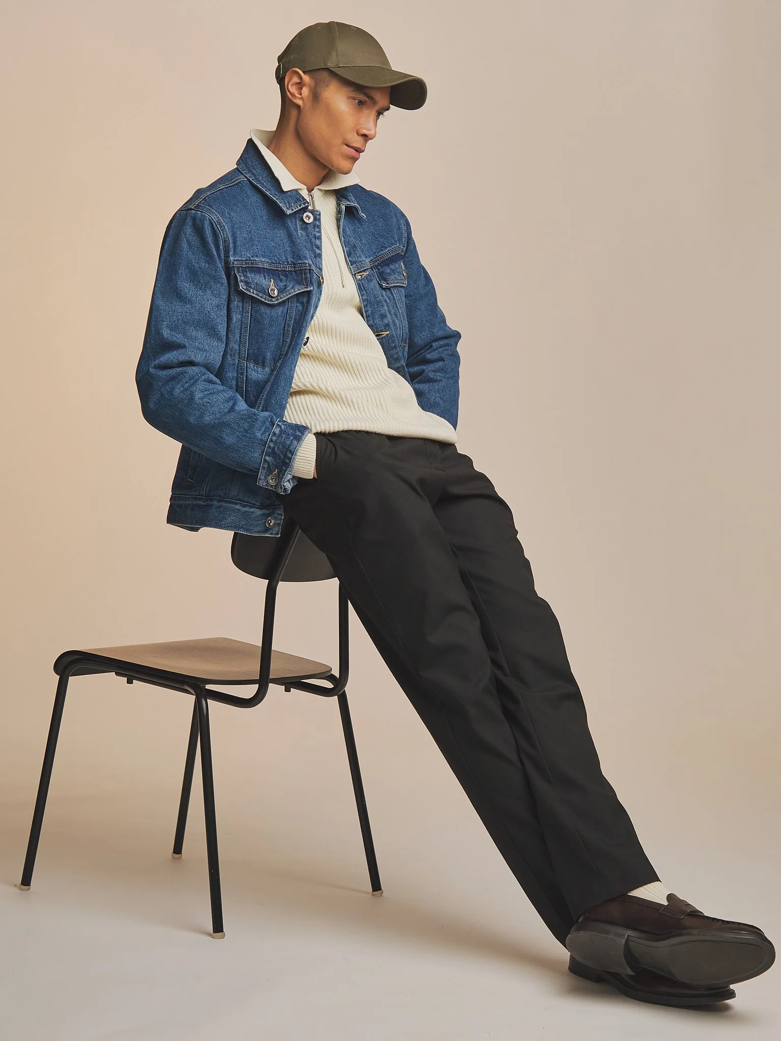 A young man sitting on a black chair, wearing a green cap, denim jacket, cream sweater, black pants, and black shoes with platform soles against a plain beige background.