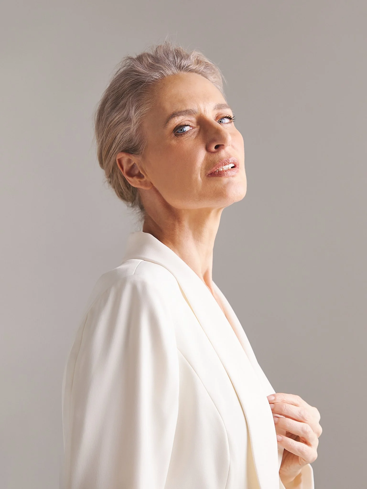A portrait of a mature woman with short, gray hair, blue eyes, wearing a white blazer, against a plain, light gray background.