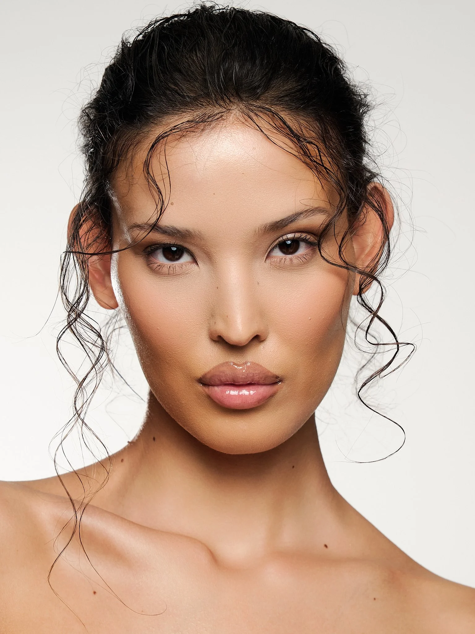 Close-up of a woman's face and upper shoulders with wet, curly black hair and a smooth complexion against a plain white background.