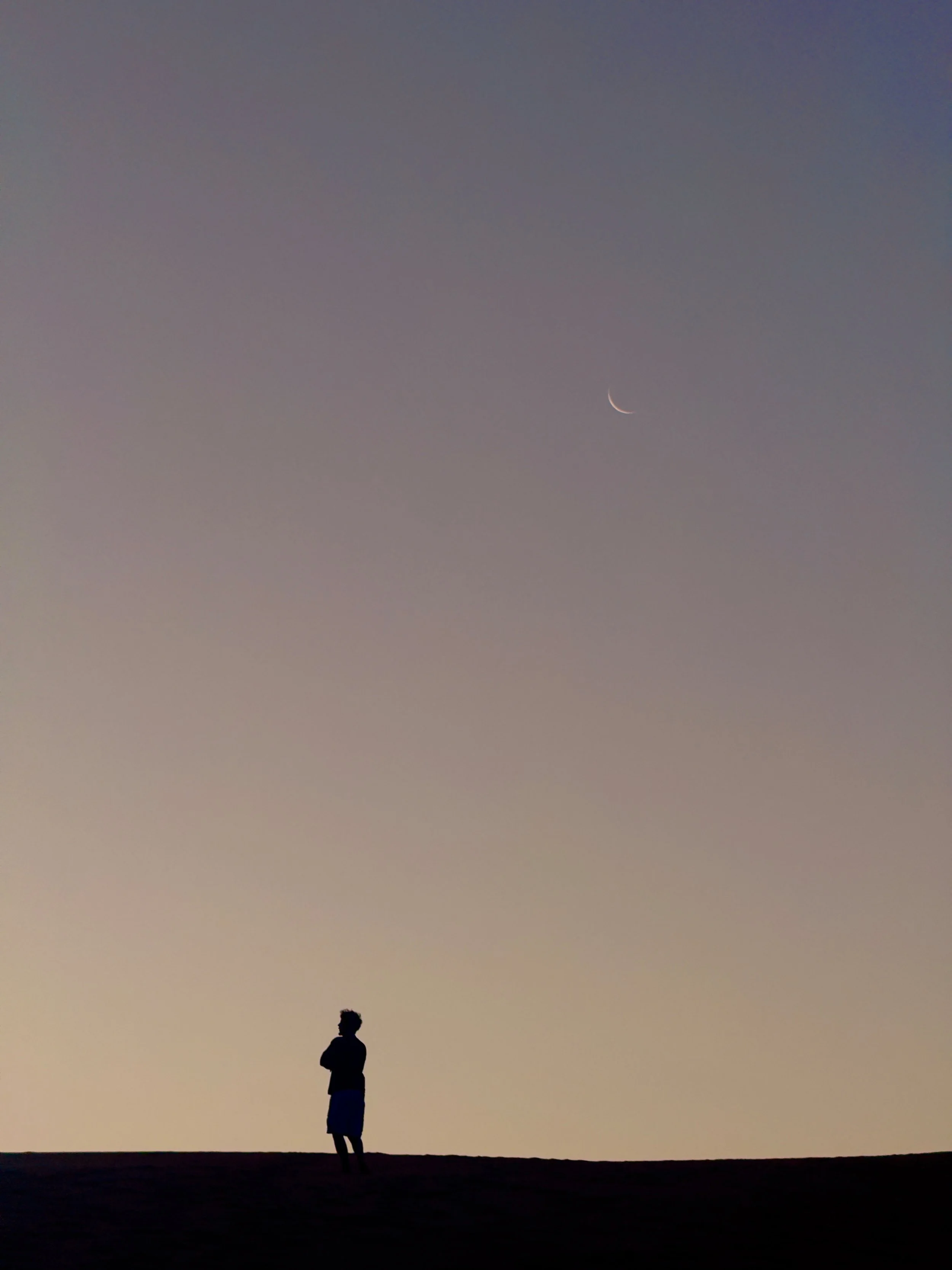 Silhouette of a person standing on a flat surface at sunset or dusk, with a gradient sky and a thin crescent moon visible in the sky.