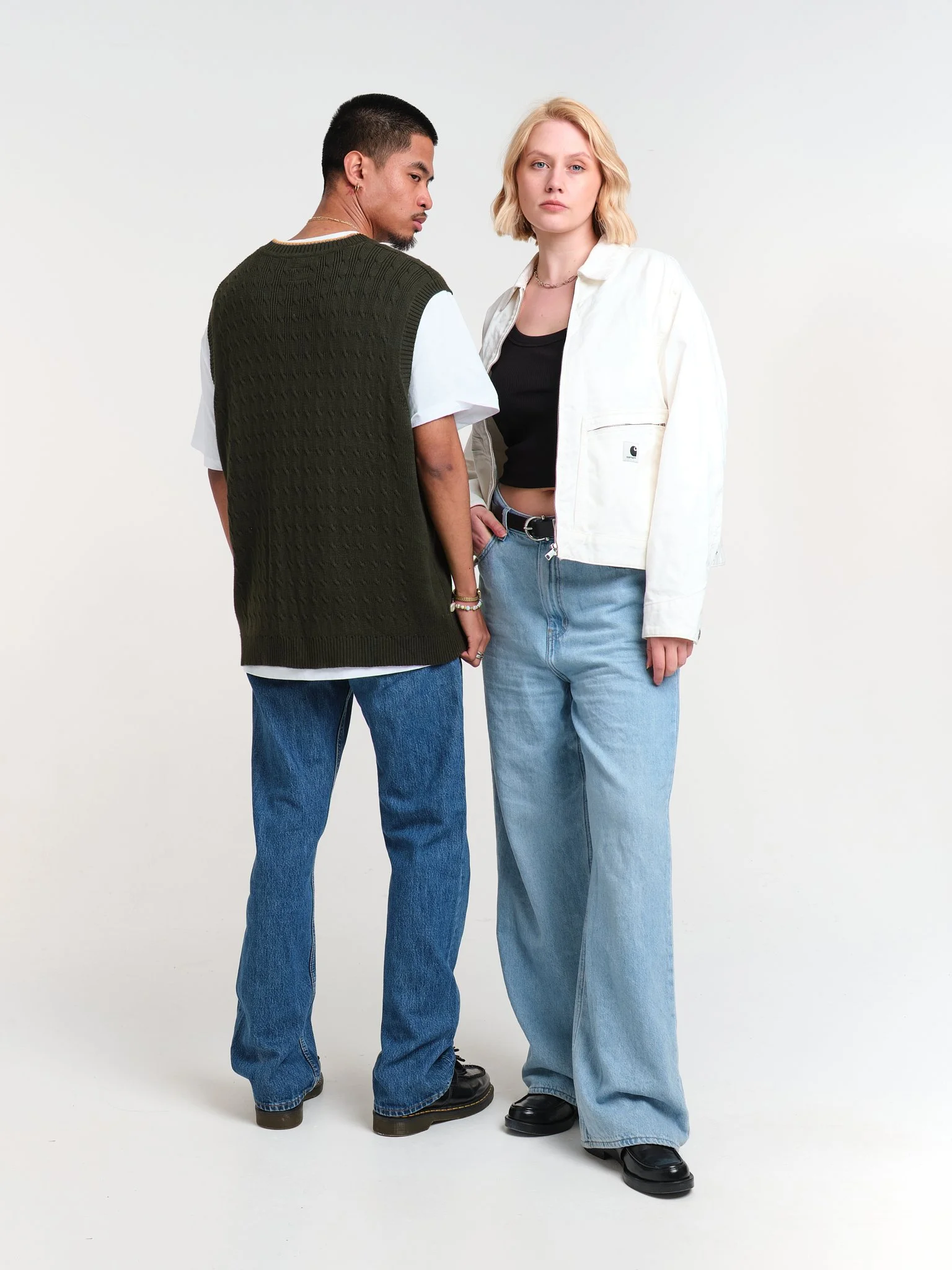 A man and woman standing against a white wall, with the woman looking directly at the camera and the man looking to the side.