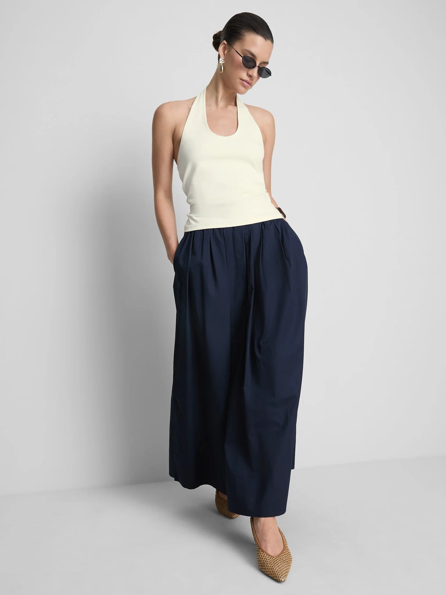 Woman in cream halter top, navy wide-leg pants, tan heels, and sunglasses against a plain gray background.