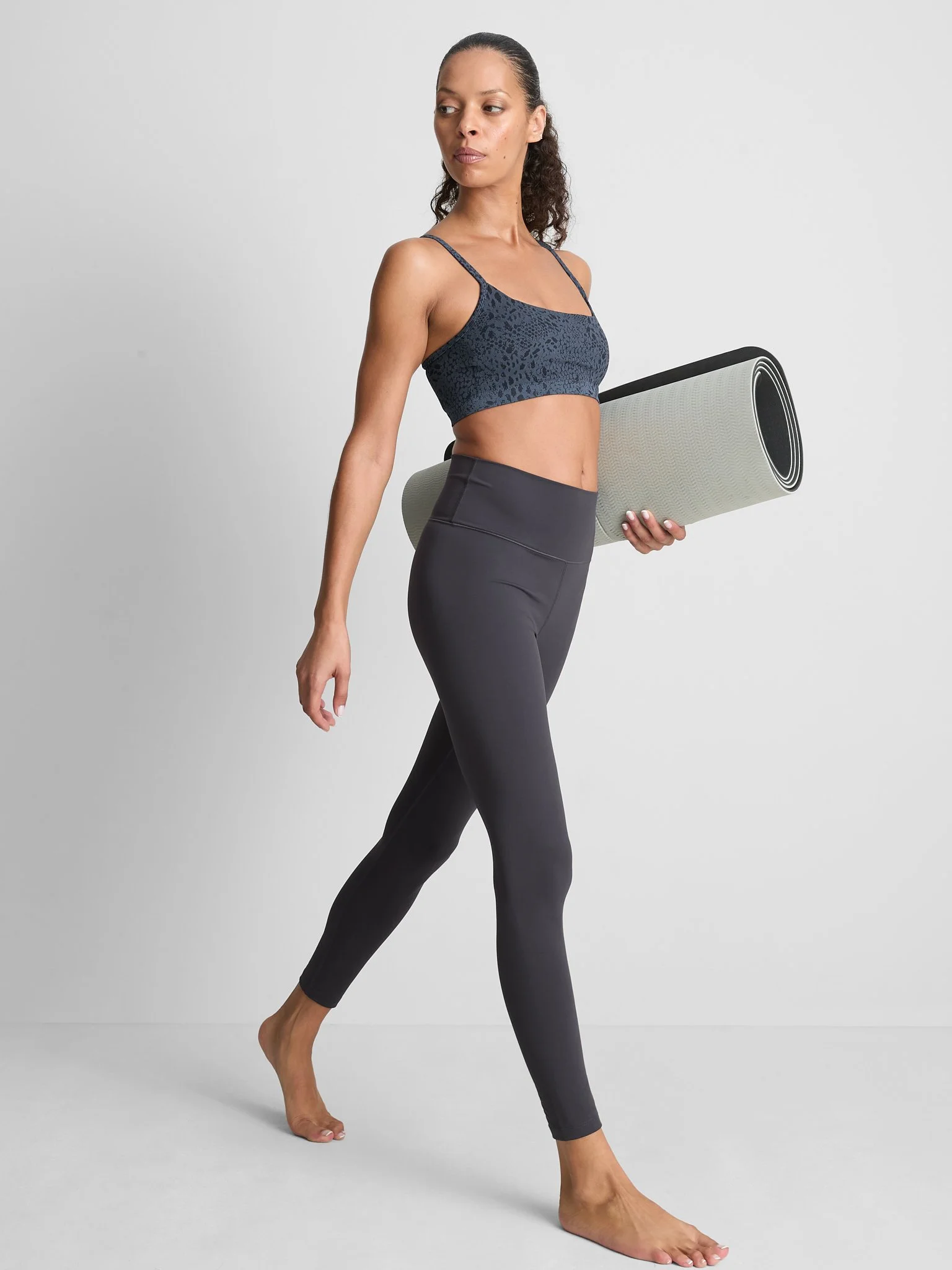 A woman in black leggings and a sports bra holding a rolled yoga mat, walking barefoot in a neutral setting.