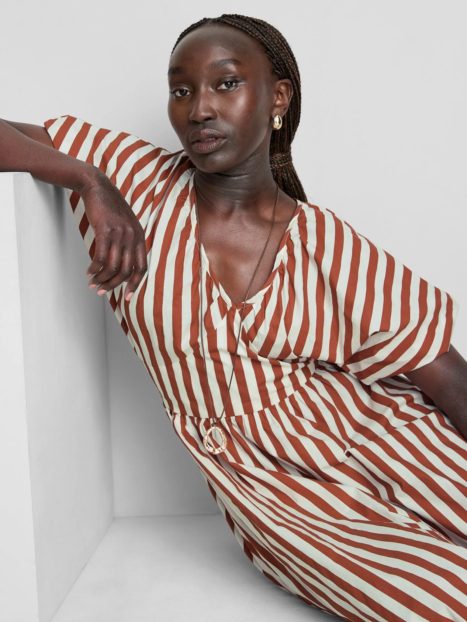 A woman with dark skin and braided hair, wearing a striped brown and white dress, is leaning against a white wall with one arm resting on it, looking at the camera with a neutral expression.
