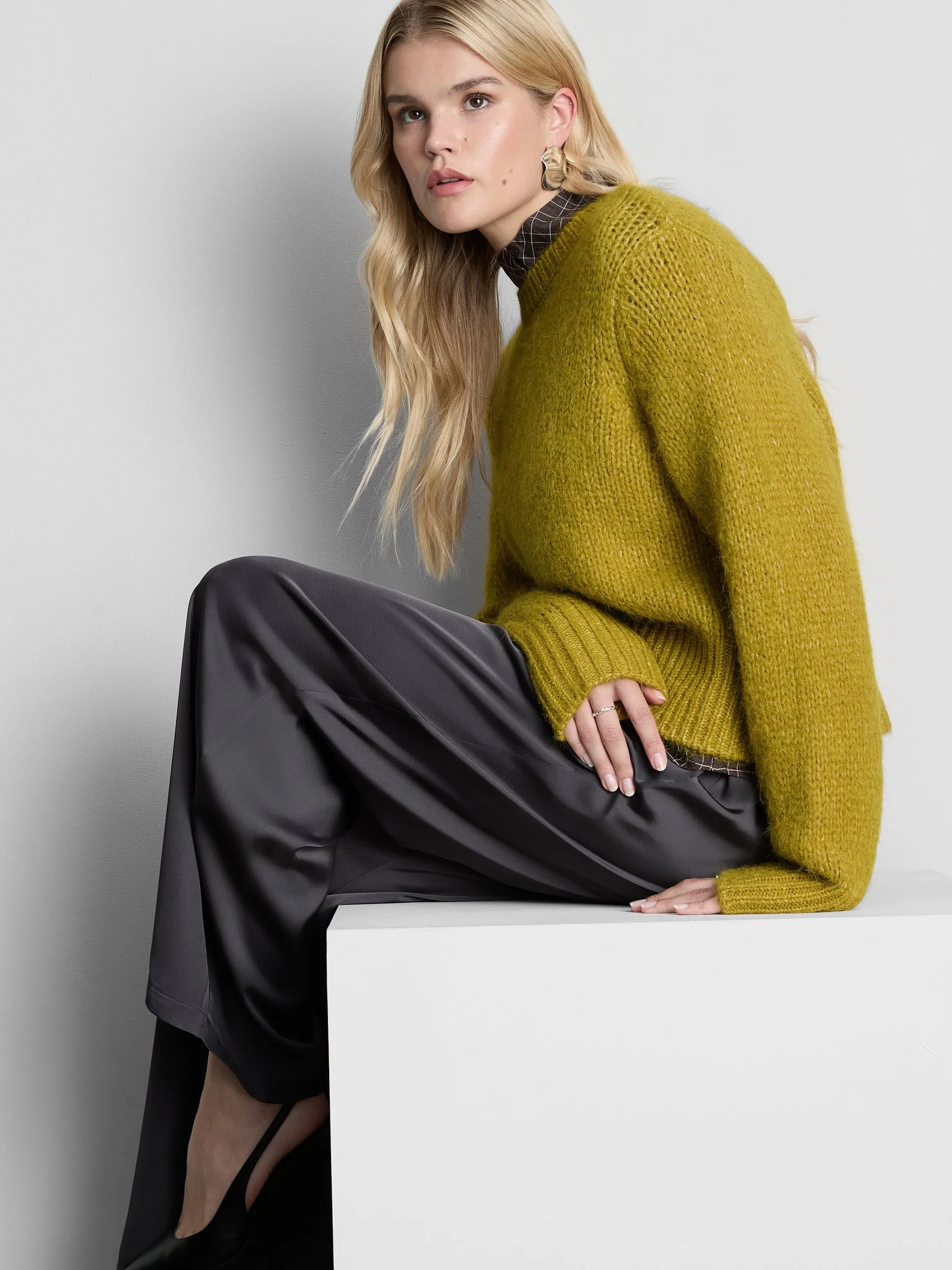 A woman with long blonde hair, wearing a mustard yellow knit sweater over a dark checkered top, black shiny pants, and black high heels. She is sitting on a white block against a plain white background, with her left arm resting on her knee and her r