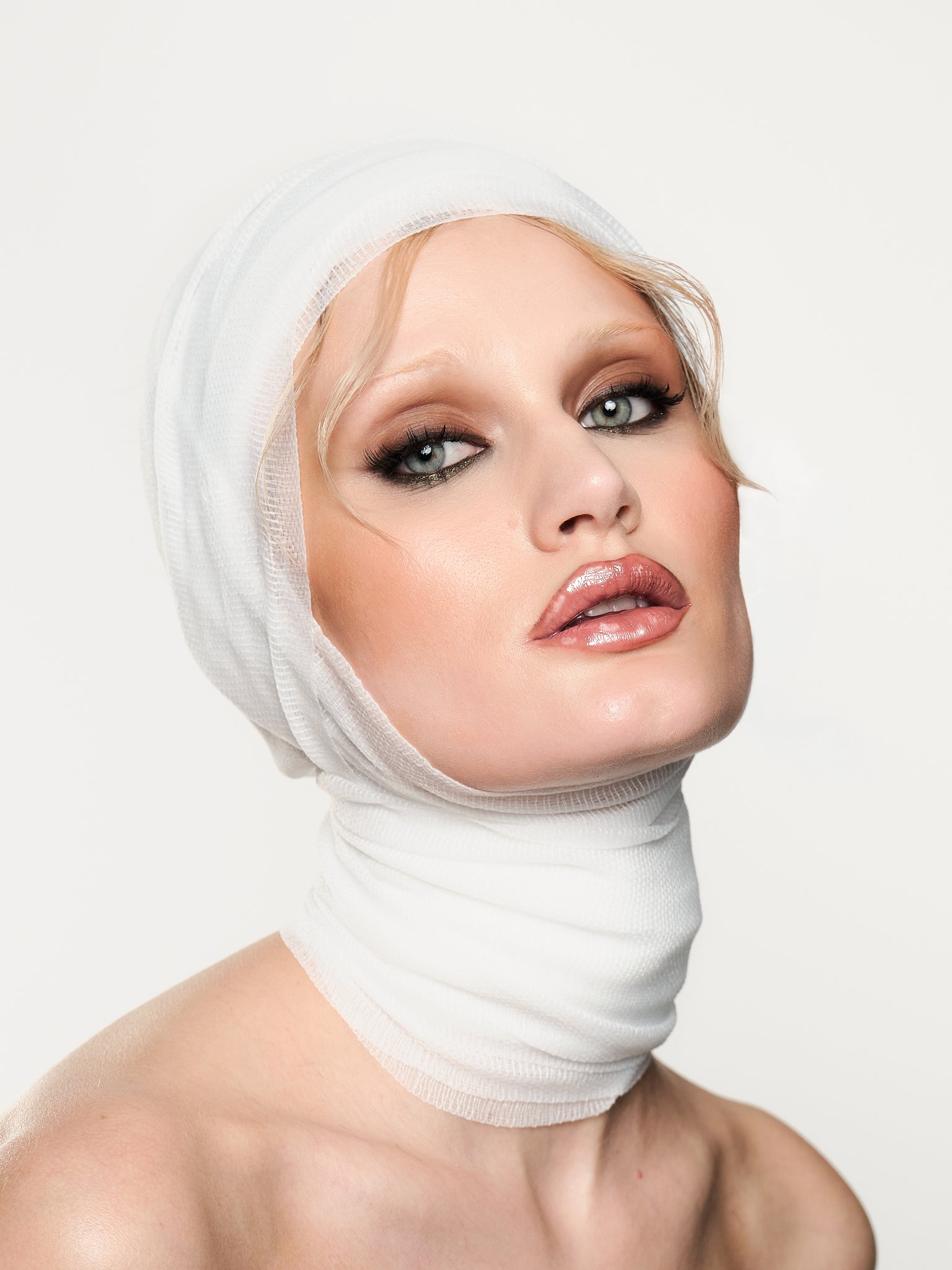A woman wearing a white head covering and white neck wrap, with makeup and glossy lips, looking at the camera against a plain background.