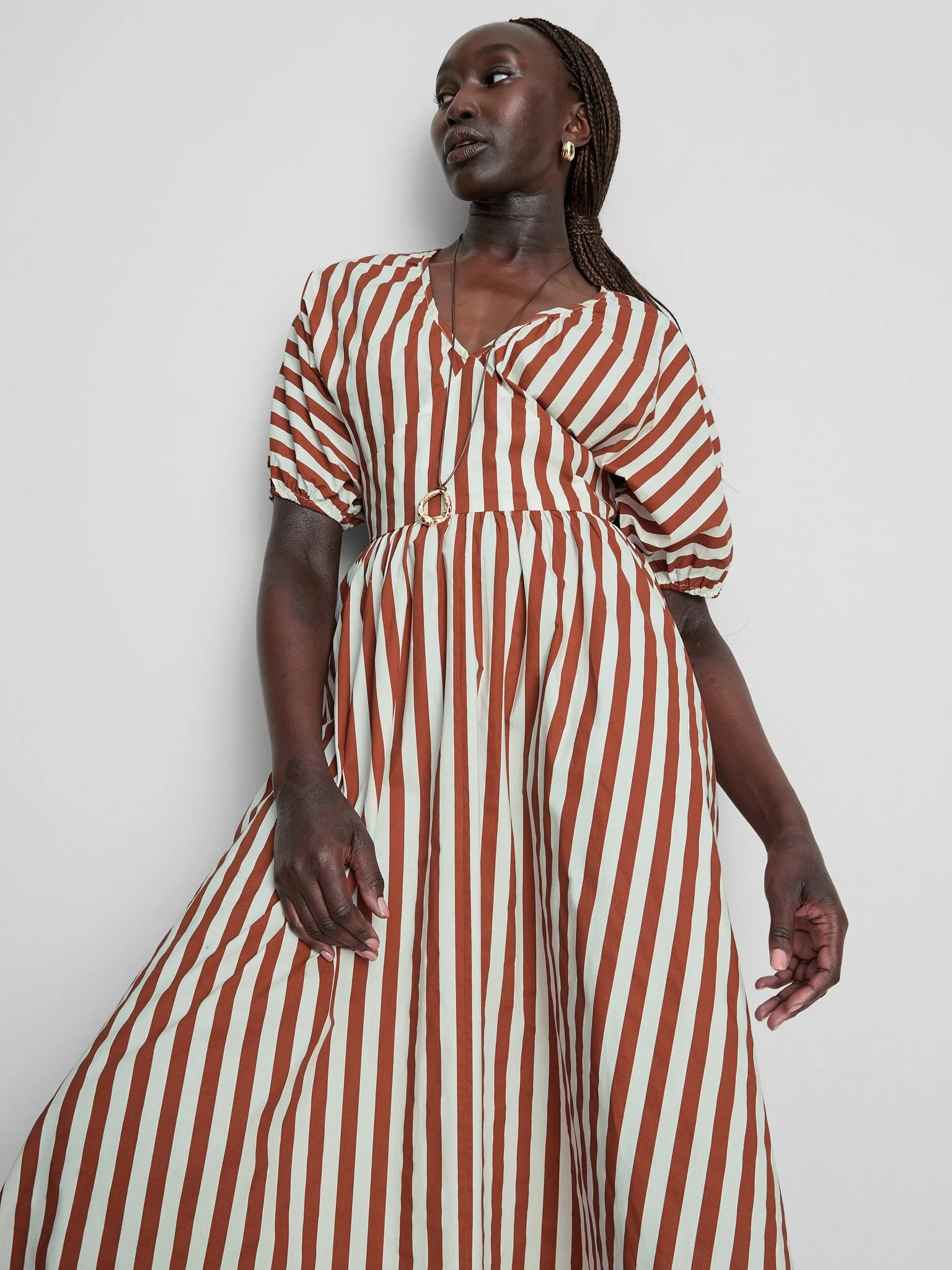 A woman with dark skin and braided hair is wearing a striped brown and white dress with puffy sleeves, accessorized with gold jewelry, standing against a plain white wall.