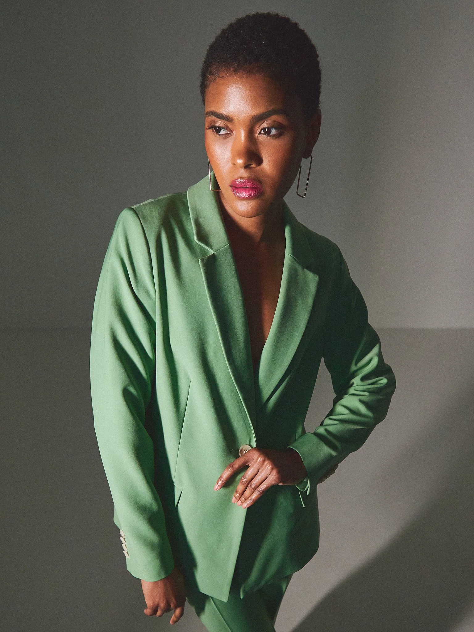 A woman with short curly hair wearing a green satin blazer and matching pants, standing against a plain gray background.