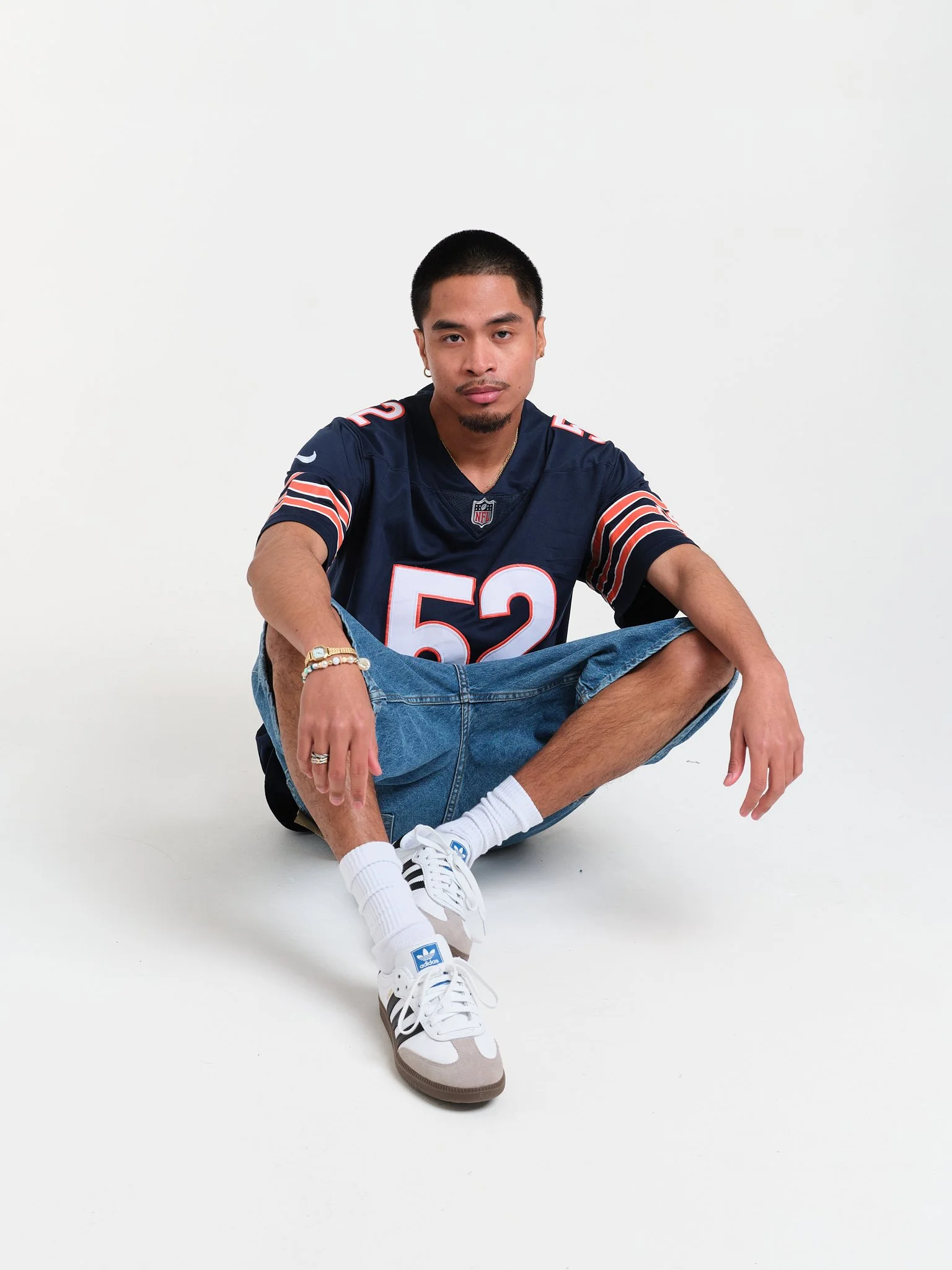 Young man sitting cross-legged on the floor in front of a white background, wearing a navy blue NFL sports jersey, jeans, white sneakers, and accessorized with jewelry.
