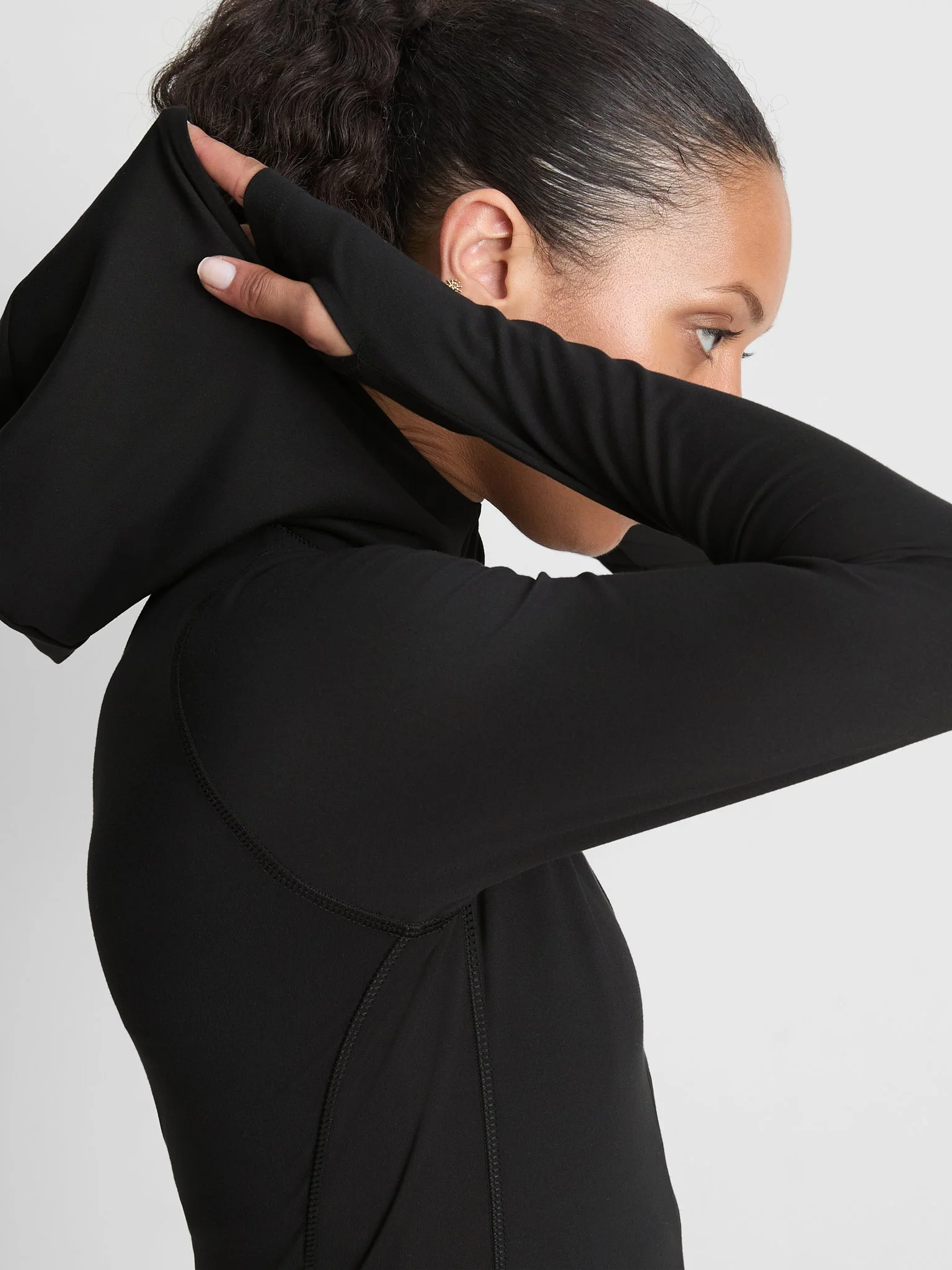 A woman with dark hair styled in a ponytail, dressed in black activewear, stretching her arm across her face against a plain background.