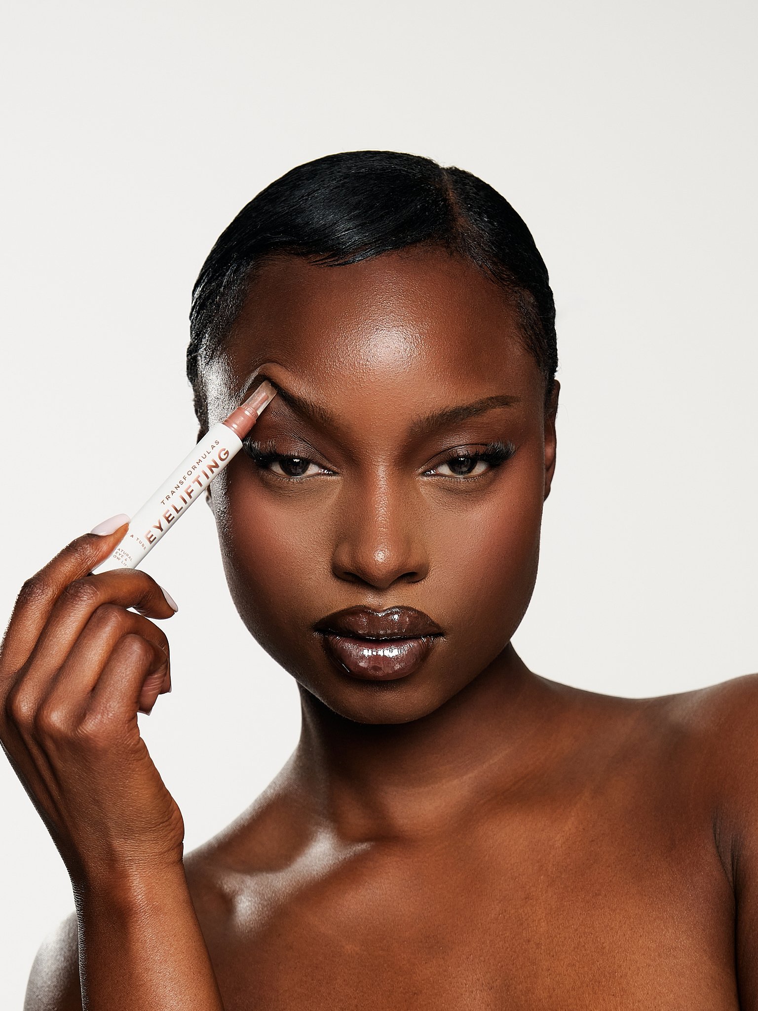Close-up of a woman with dark skin, holding a tube of eyelift product near her eye, with smooth skin and makeup, against a plain white background.