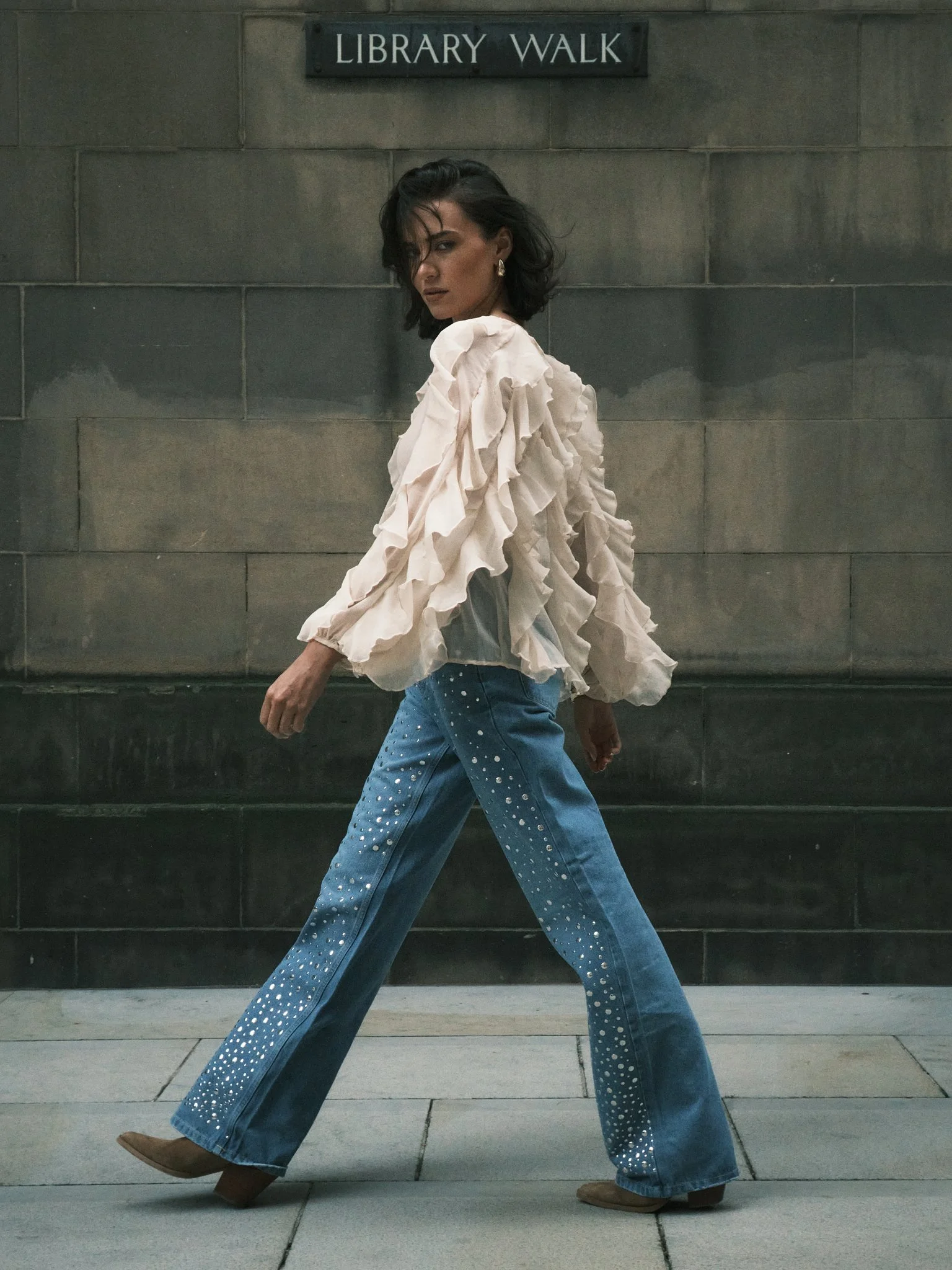 A woman walking in front of a wall with a sign that says 'Library Walk'. She has shoulder-length dark hair, wears gold earrings, a ruffled beige blouse, and blue jeans decorated with white rhinestones.