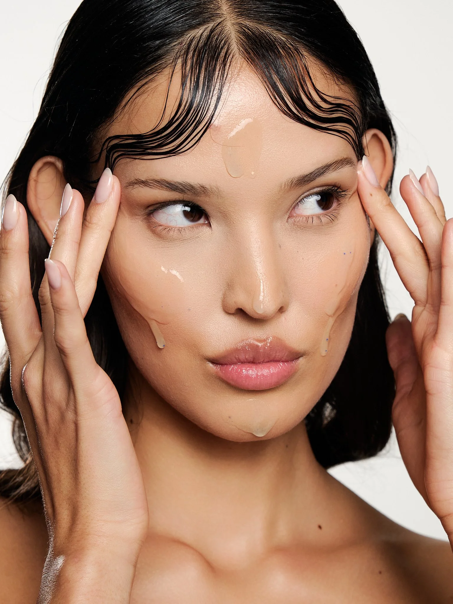 A woman with wet hair and light skin is holding her temples with both hands. She has a few drops of water on her face and appears to be in a skincare or beauty routine. The background is plain white.