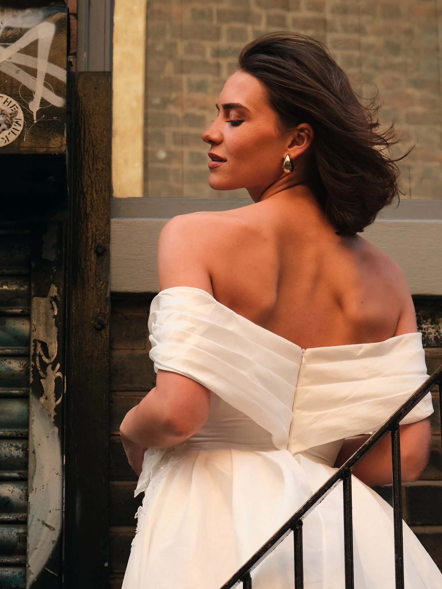 A woman with short brown hair and gold earrings wearing an off-the-shoulder white dress, sitting on a staircase with her eyes closed.